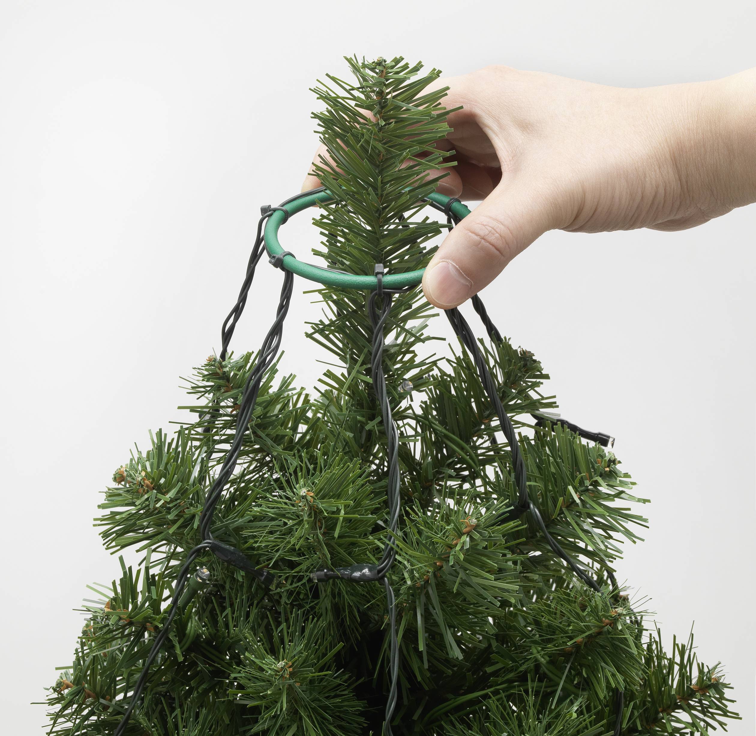Eine Hand platziert ein grünes Plastikteil oben auf einem künstlichen Weihnachtsbaum, um Lichter daran zu befestigen.