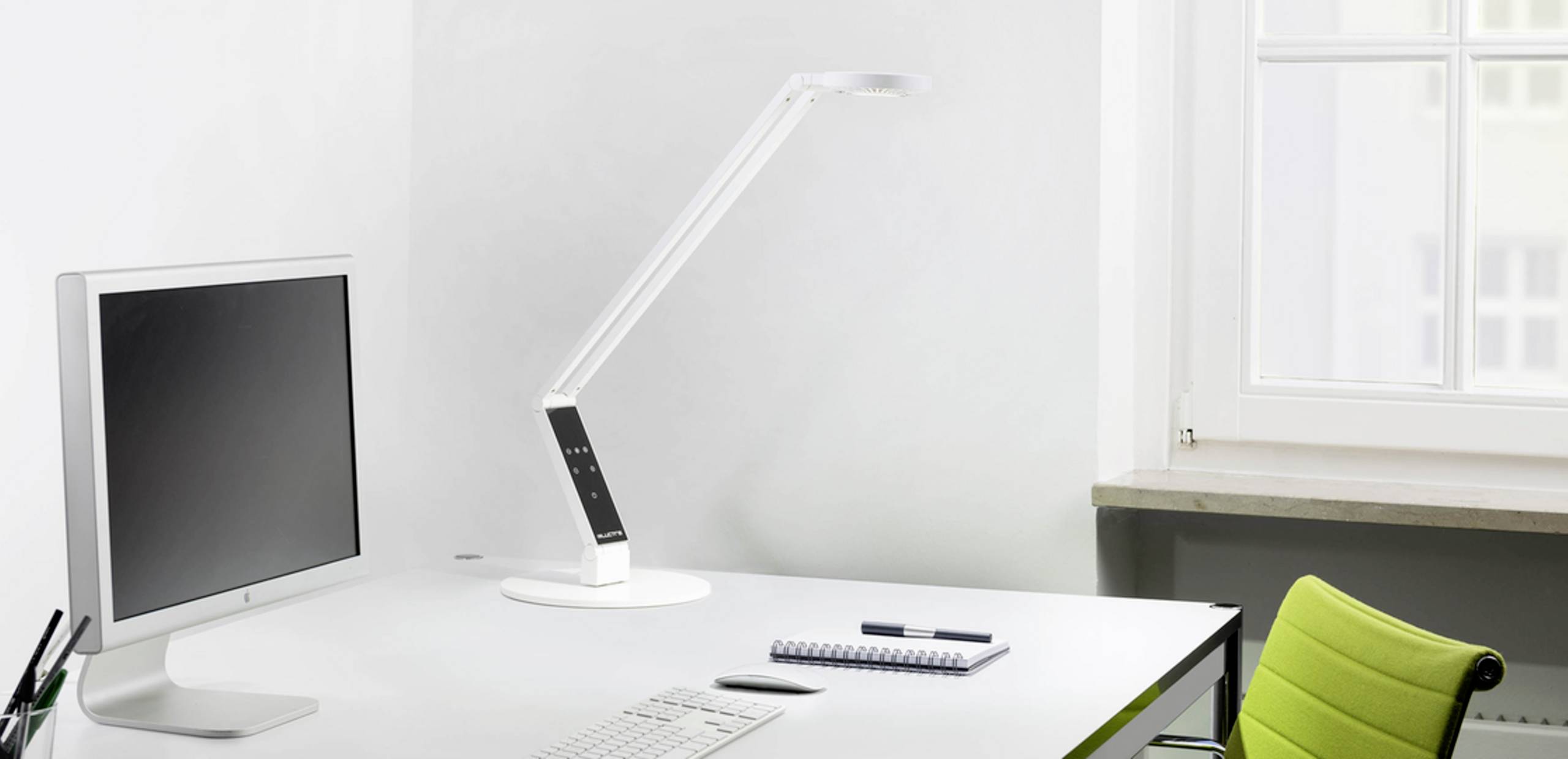 Ein moderner Schreibtisch mit einem Computerbildschirm, einer Tischlampe, einem Notizblock und einem Stift. Ein grüner Stuhl steht daneben.