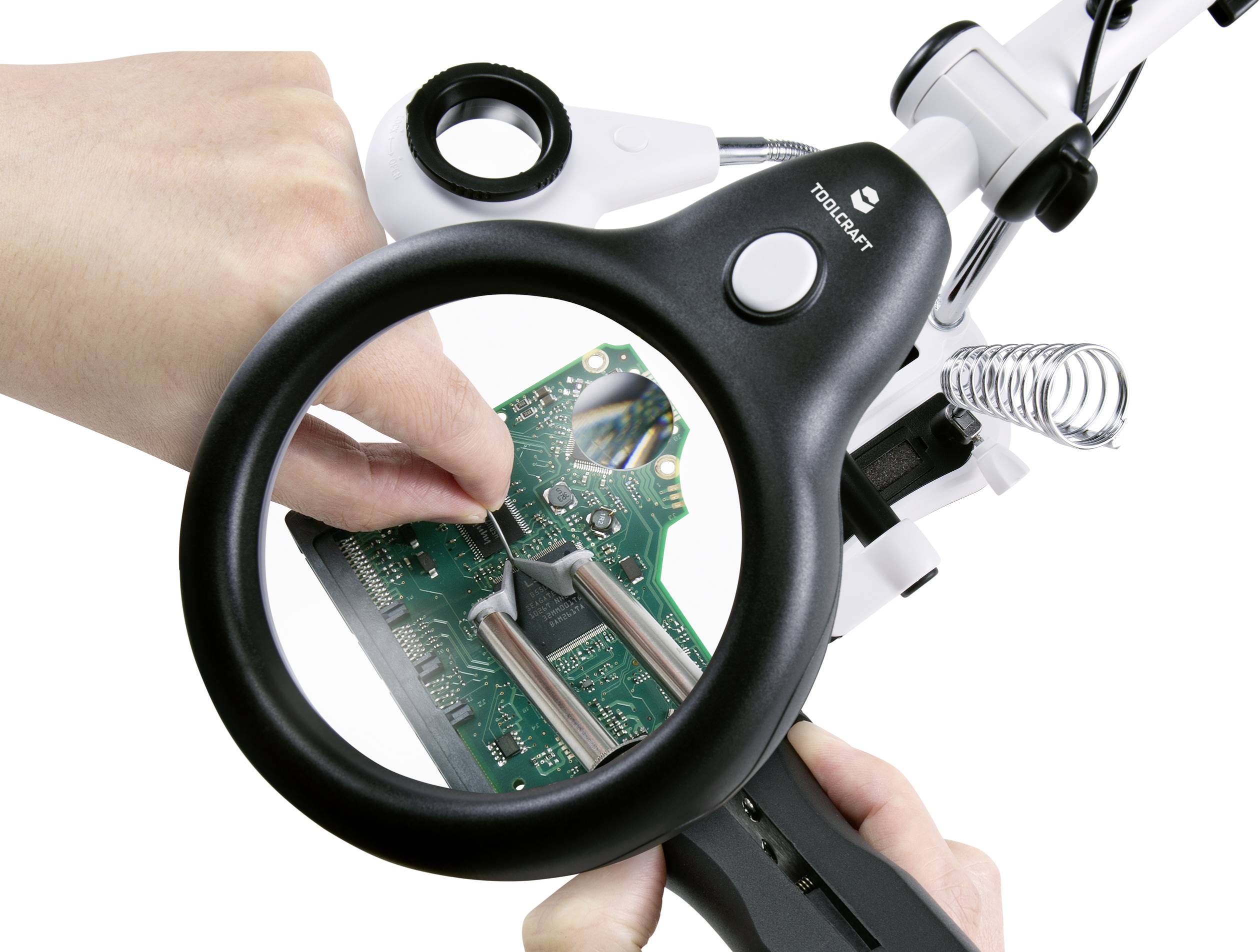 Eine Hand hält eine Lupe über eine Computerplatine, die von der anderen Hand bearbeitet wird. Die Szene zeigt Detailarbeit an Elektronik.