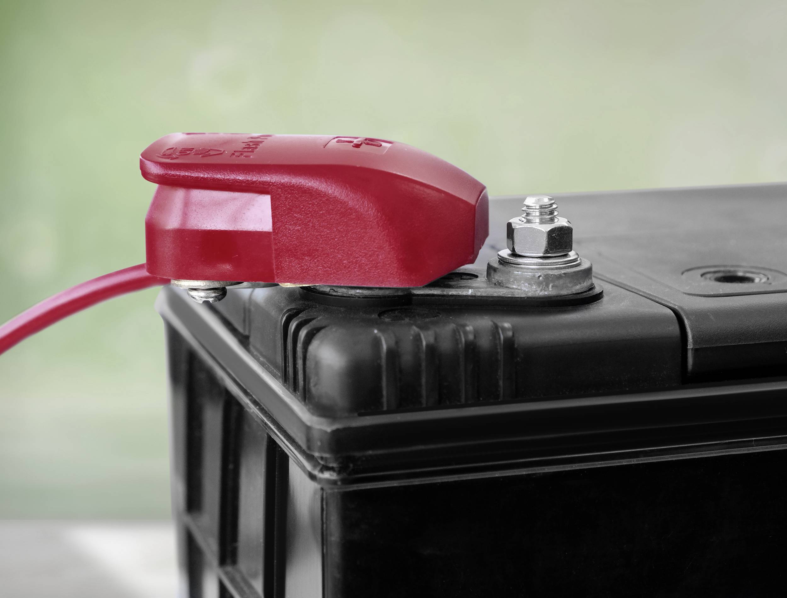 Eine Autobatterie zeigt einen roten Pluspolanschluss, der sicher befestigt ist. Der Hintergrund ist unscharf und grünlich.