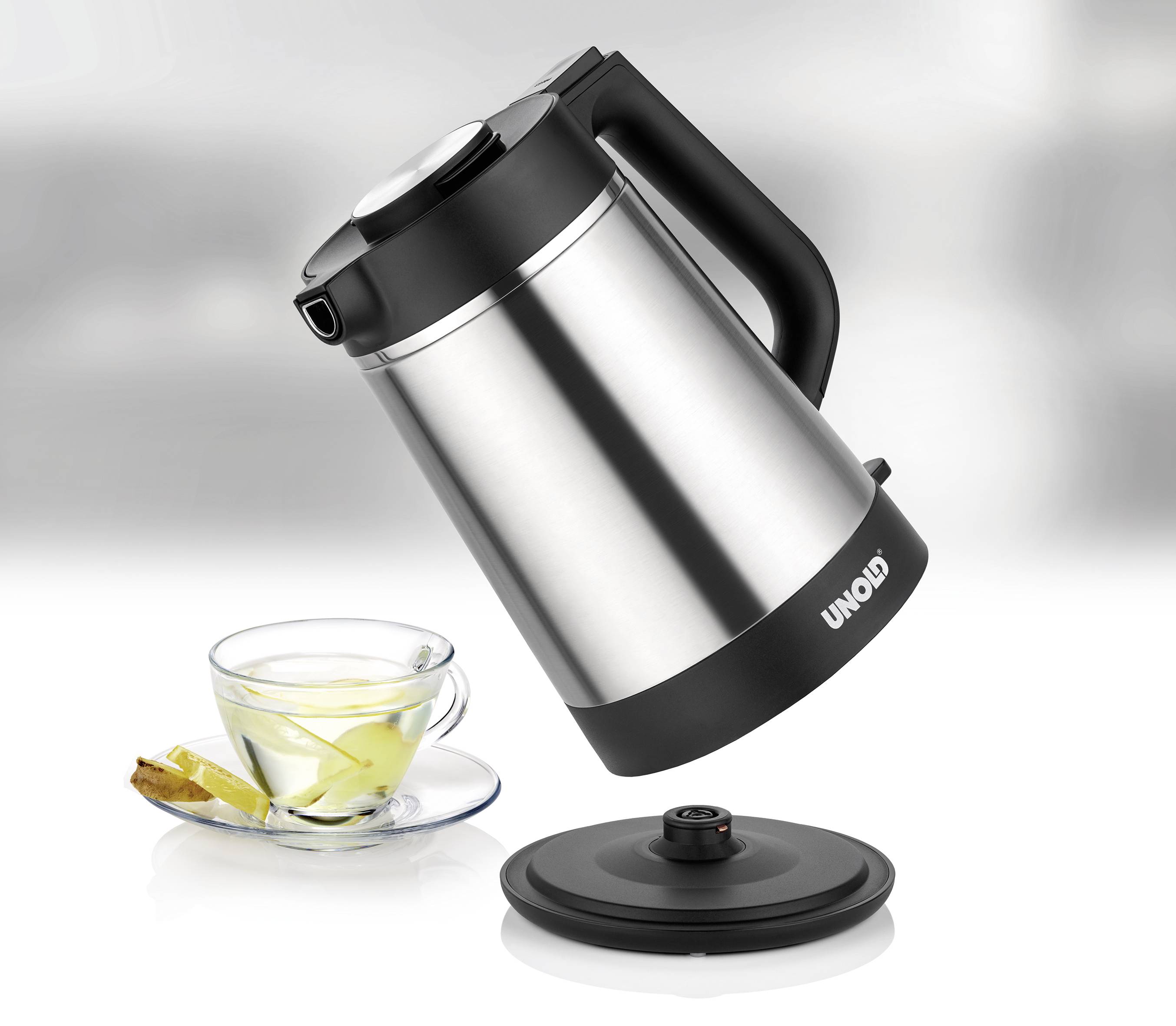 Ein Edelstahl-Wasserkocher mit schwarzem Griff wird über eine Tasse Tee mit Zitronenscheibe geleert. Daneben liegt ein schwarzer Deckel.