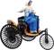 Historische Figur auf einem dreirädrigen Fahrzeug, trägt blau-weißes Gewand und sitzt auf einem Holzsitz mit schwarzen Fahrradspeichen.