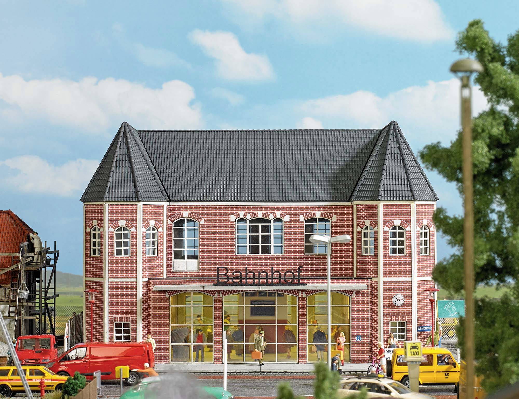 Ein Modell eines roten Backsteinbahnhofs mit Schriftzug 'Bahnhof', umgeben von Autos und Menschen, darunter ein Baum im Vordergrund.