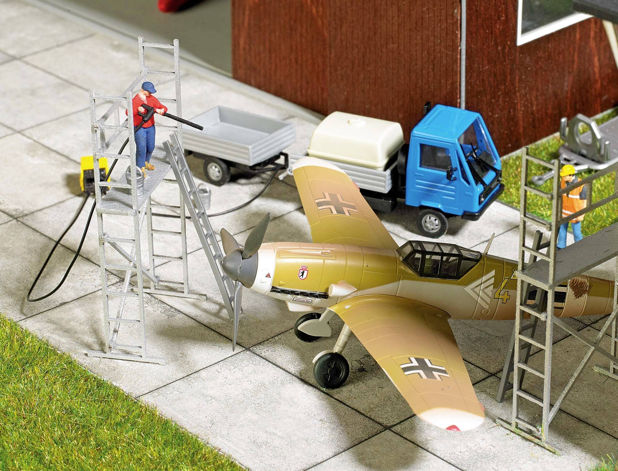 Ein Modellflugzeug wird gewartet. Zwei Männer arbeiten auf Leitern daneben. Im Hintergrund stehen ein blauer Transporter und ein Anhänger.