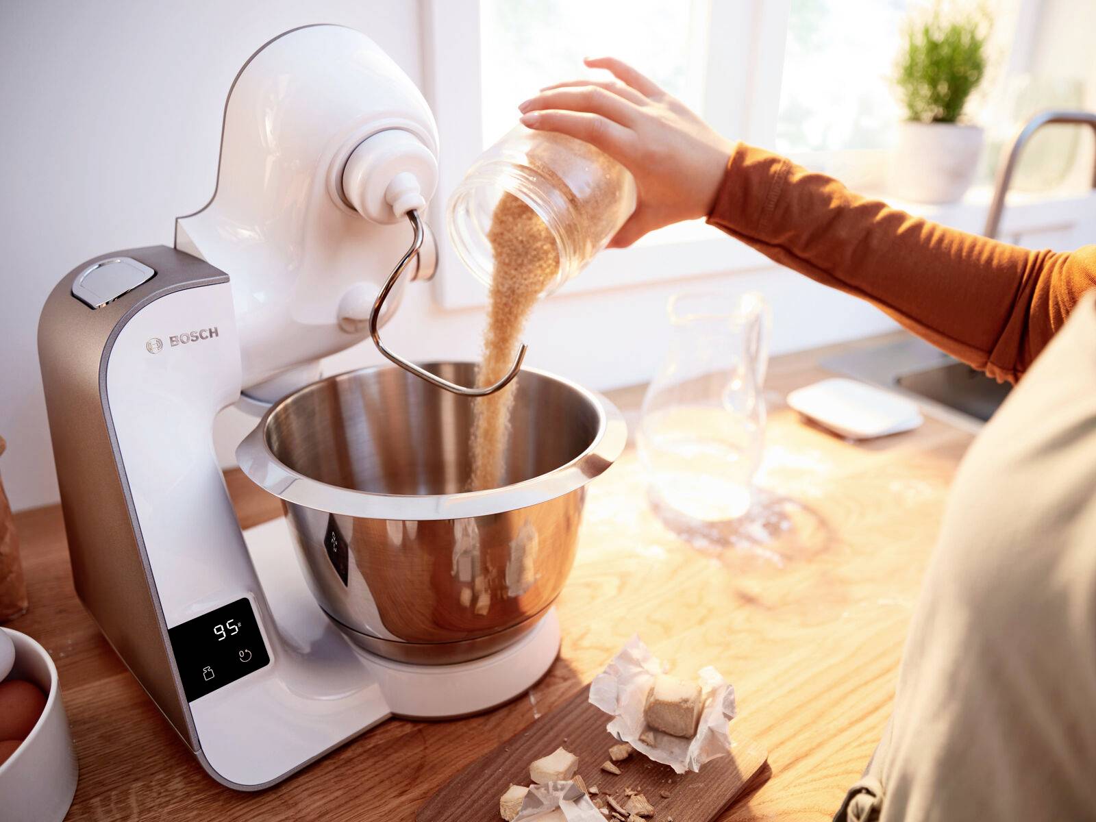Eine Person gießt Zucker in eine laufende Küchenmaschine auf einem Holztisch. Im Hintergrund sind ein Fenster und Gewürze zu sehen.