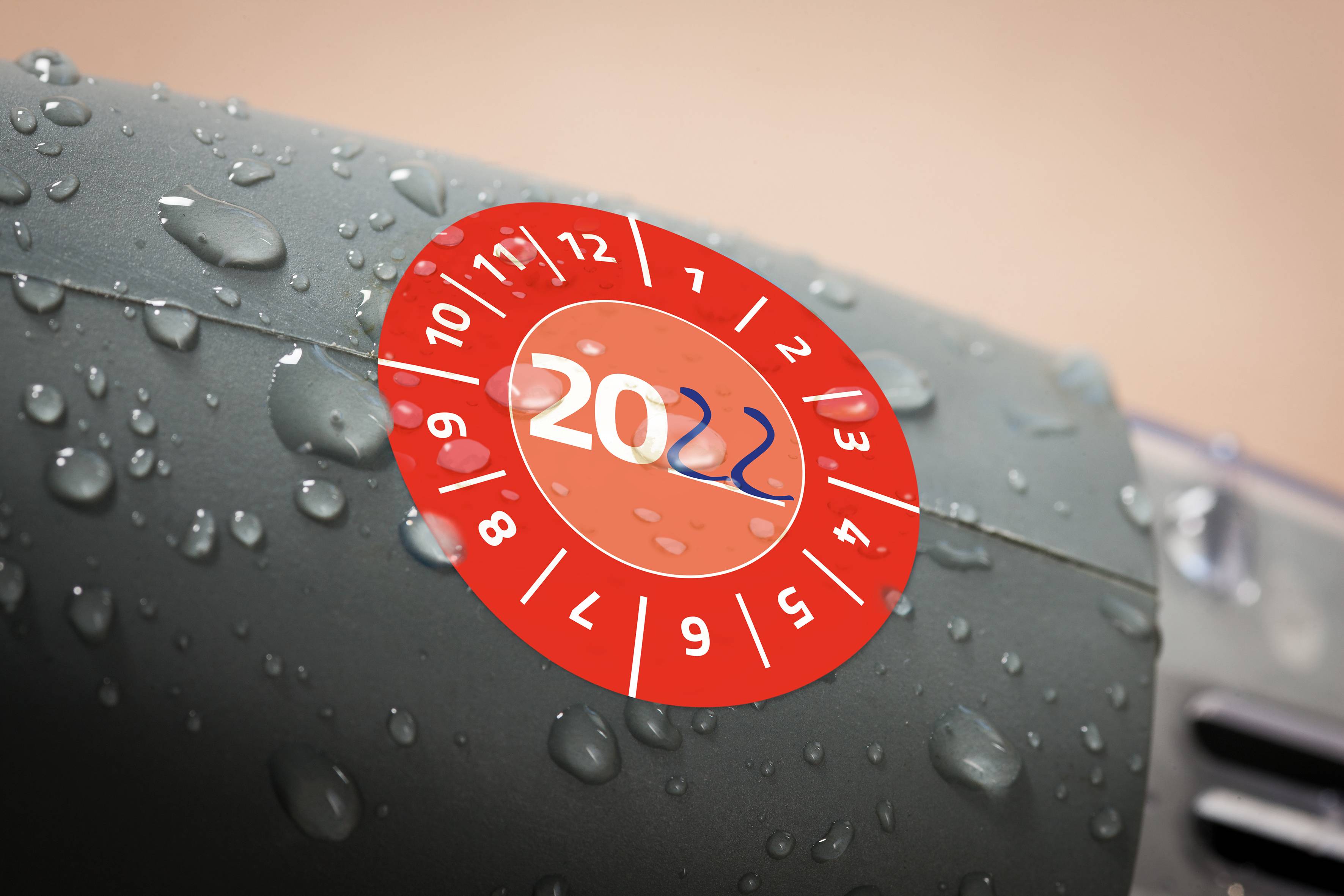 'Rote Prüfplakette mit dem Jahr 2022 für TÜV-Untersuchung auf regennasser Oberfläche eines Fahrzeugs, zeigt Frist bis November.'