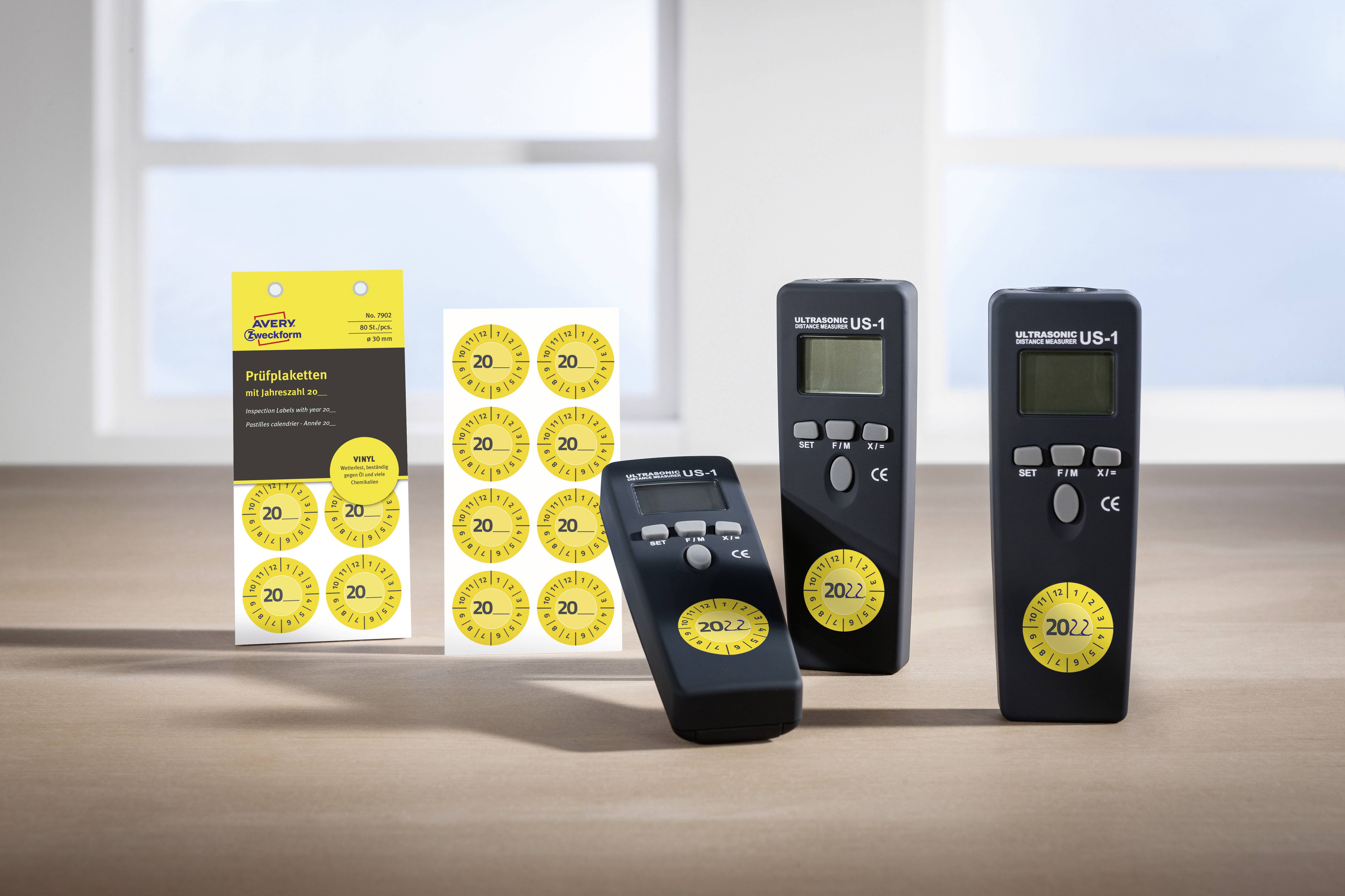 Drei schwarze Geräte mit Bildschirmen stehen auf einem Tisch neben einer Karte mit gelben Prüfplaketten. Ein Fenster im Hintergrund.