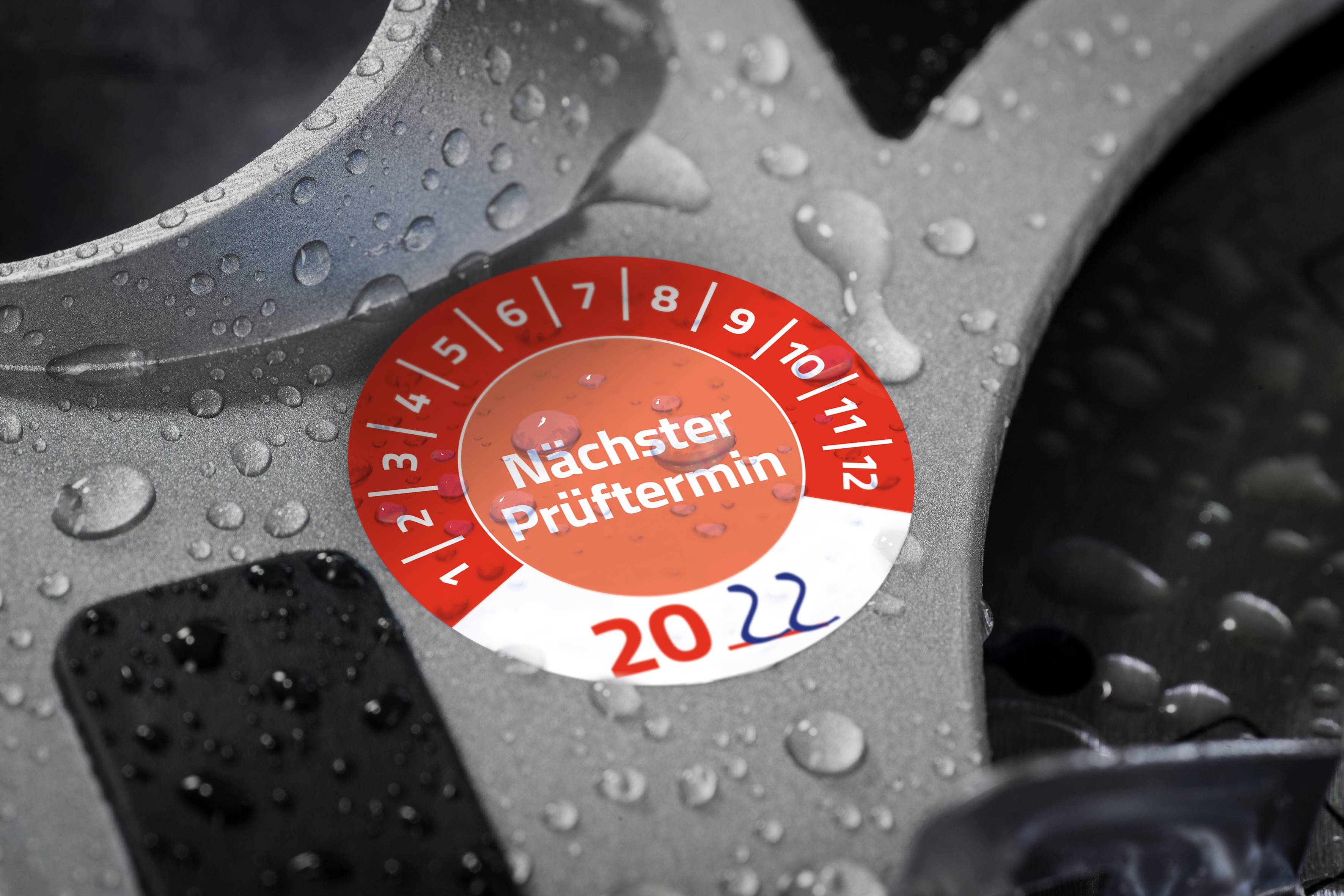 Prüfplakette 'Nächster Prüftermin' auf Autoreifen mit Jahresangabe '2022', umgeben von Wassertropfen.