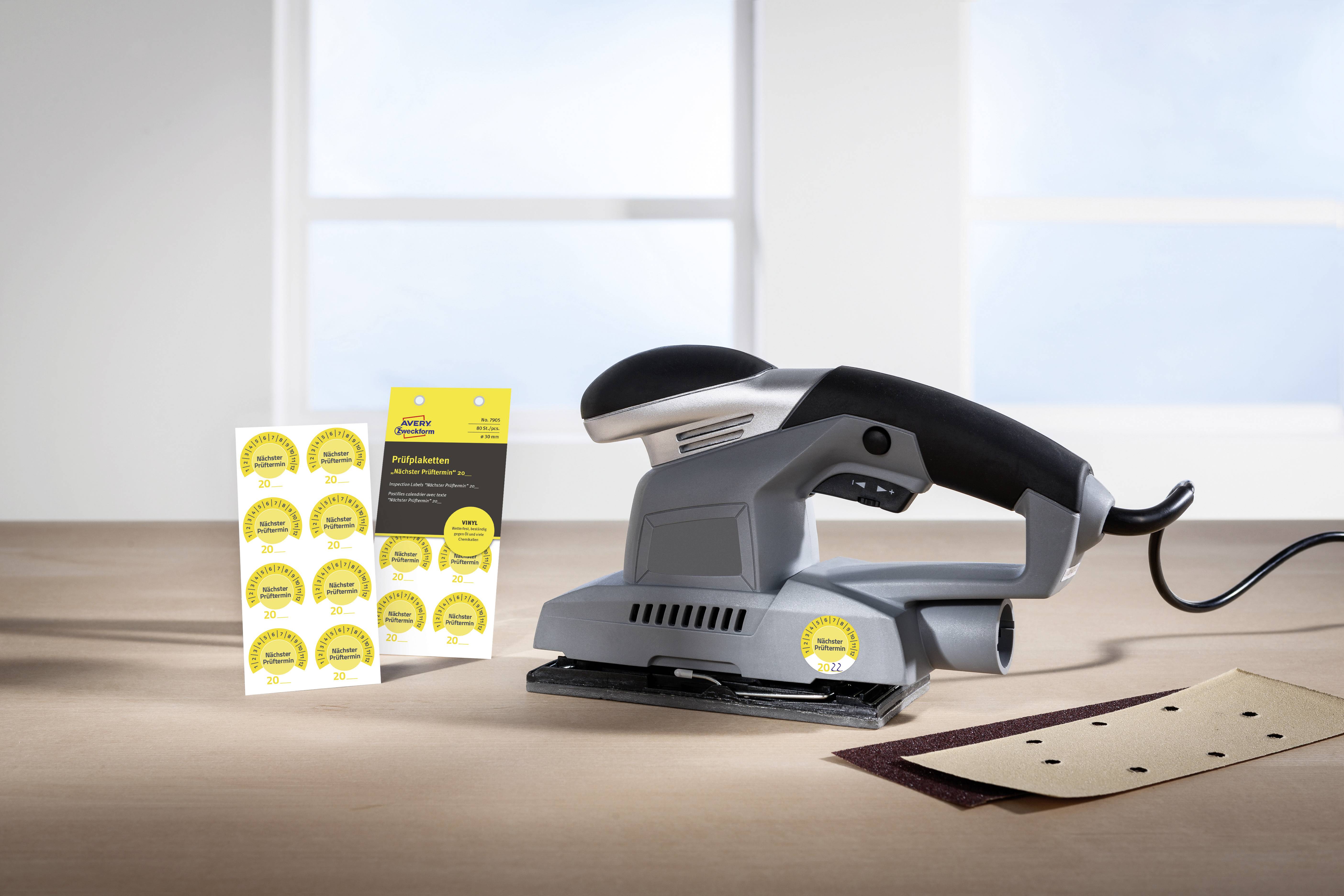 'Handschleifgerät auf Tisch mit Schleifpapier in neutraler Umgebung; Verpackung mit Ersatz-Klettpads daneben.'