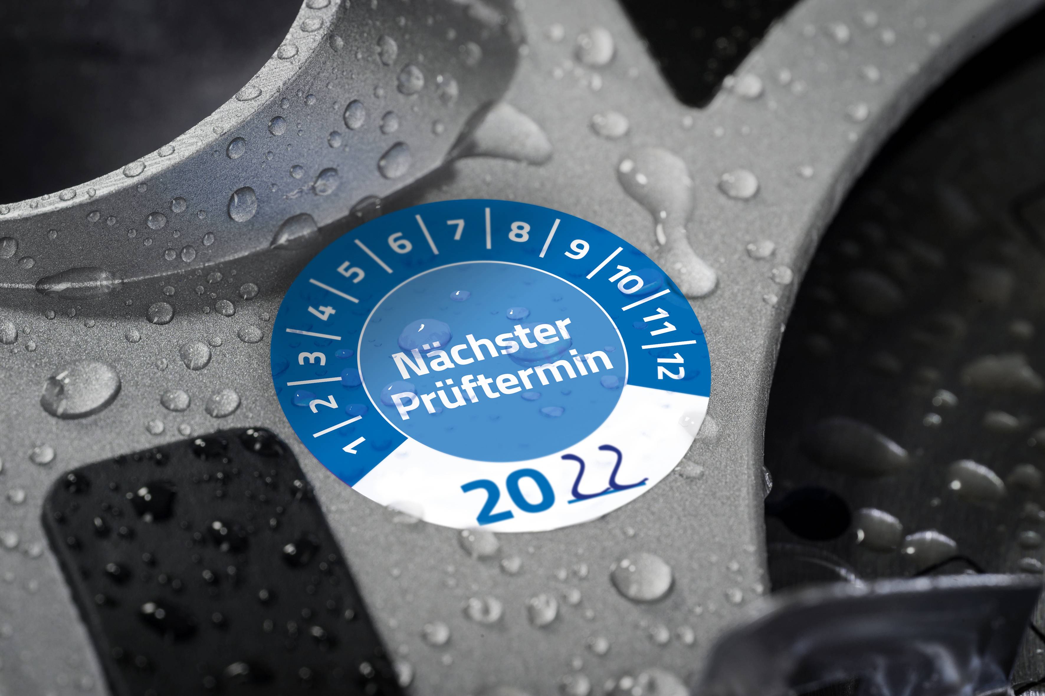 Auf einem nassen Autoteil befindet sich eine blaue Plakette mit der Aufschrift 'Nächster Prüftermin' und '2022', umgeben von Monatszahlen.