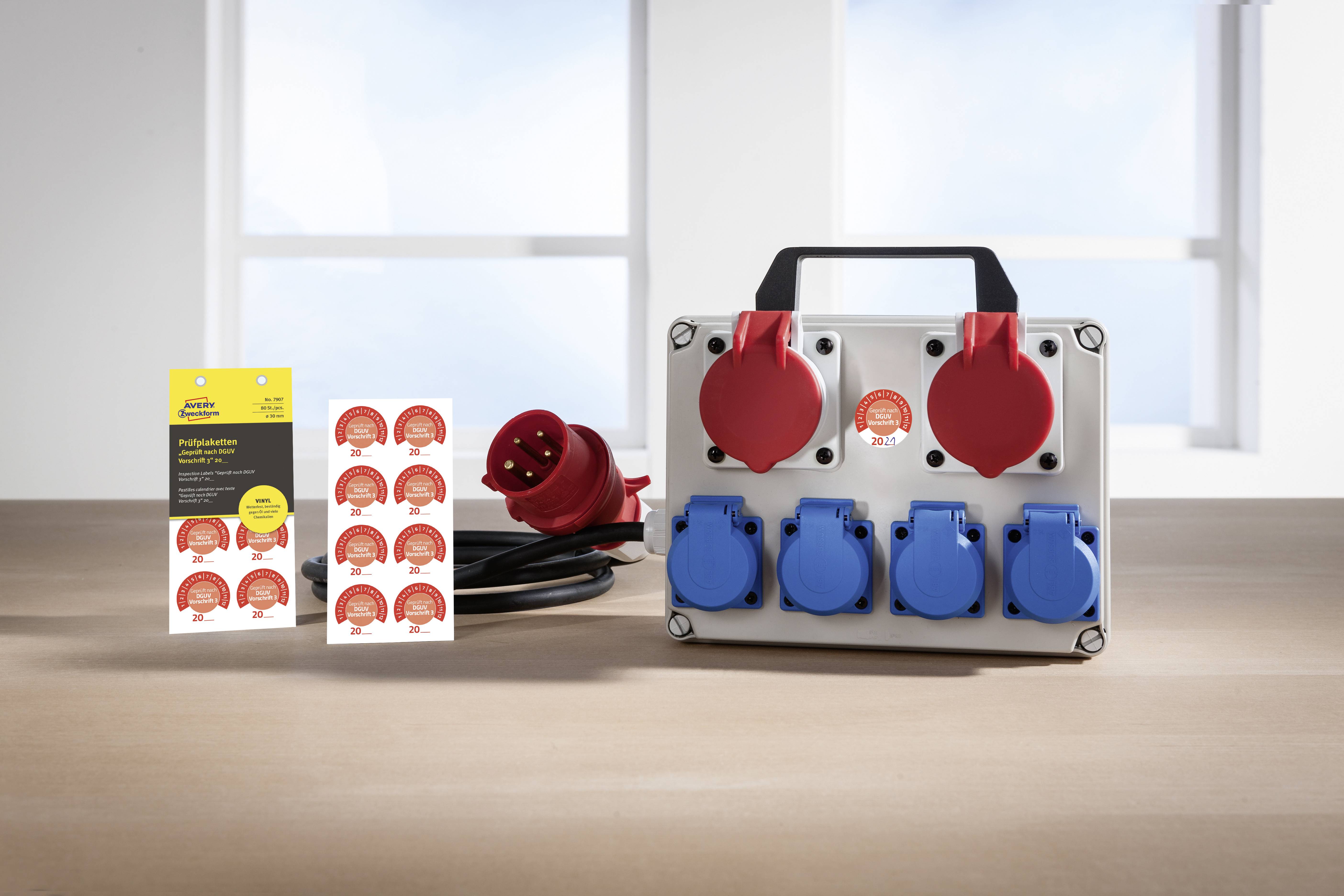 Das Bild zeigt eine tragbare Steckdosenleiste mit drei roten und vier blauen Steckdosen. Daneben liegen Aufkleber und eine Betriebsanleitung.