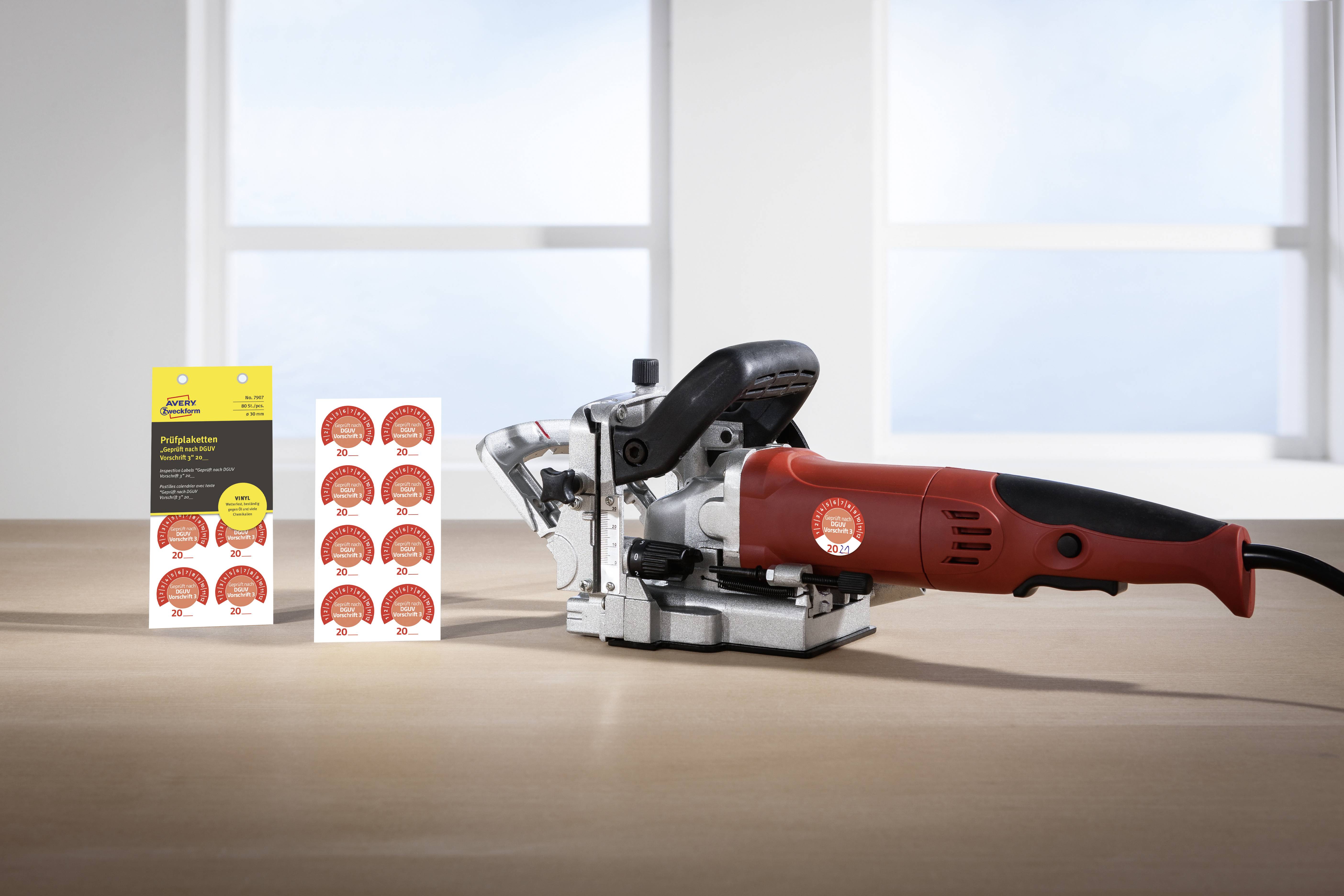 Eine rote Handfräse steht auf einem Tisch neben einer Packung Holzdübel. Im Hintergrund sind zwei helle Fenster zu sehen.