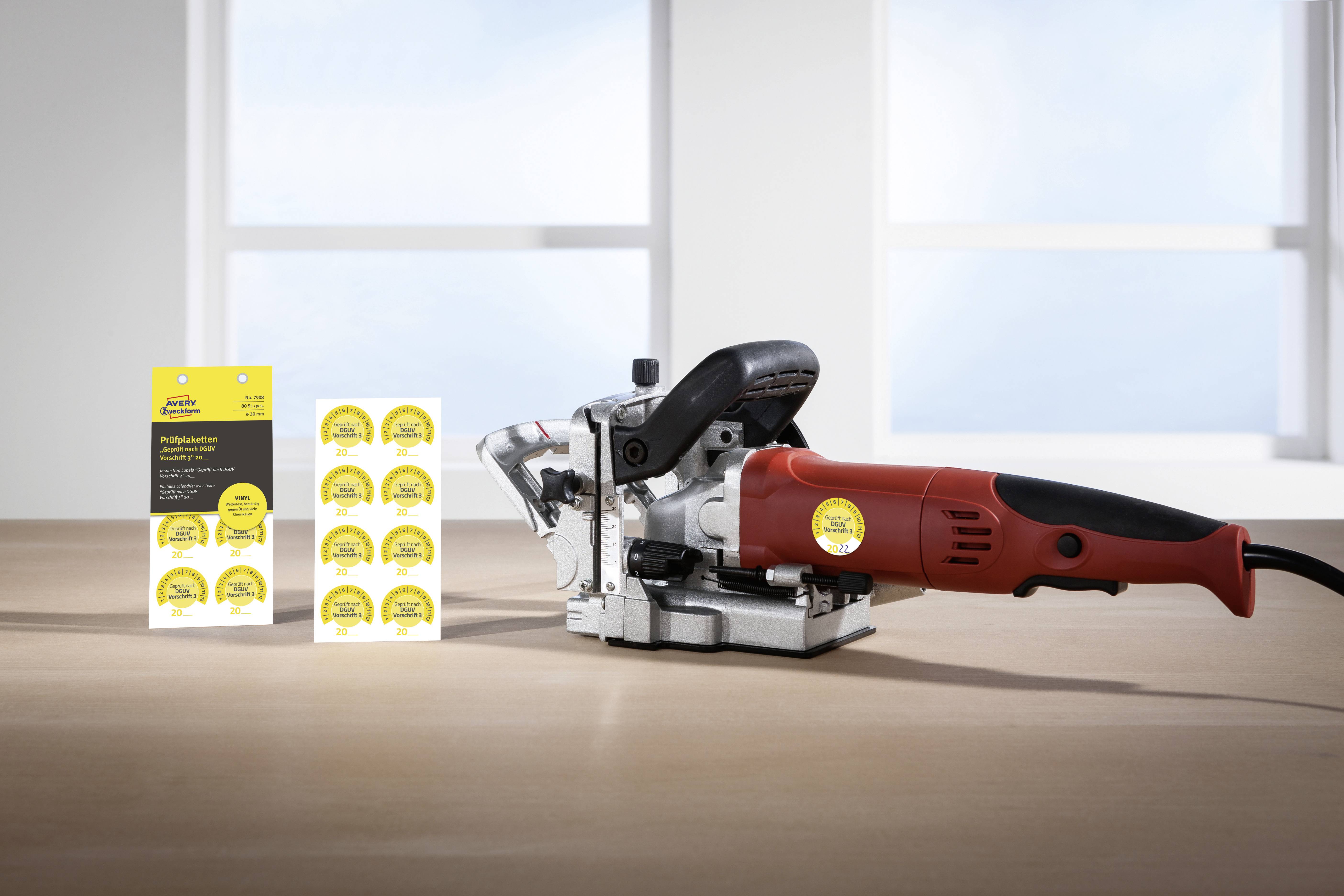 Eine rote elektrische Handfräse steht auf einem Tisch. Daneben sind mehrere gelbe Aufkleber mit schwarzem Text zu sehen. Im Hintergrund ein Fenster.