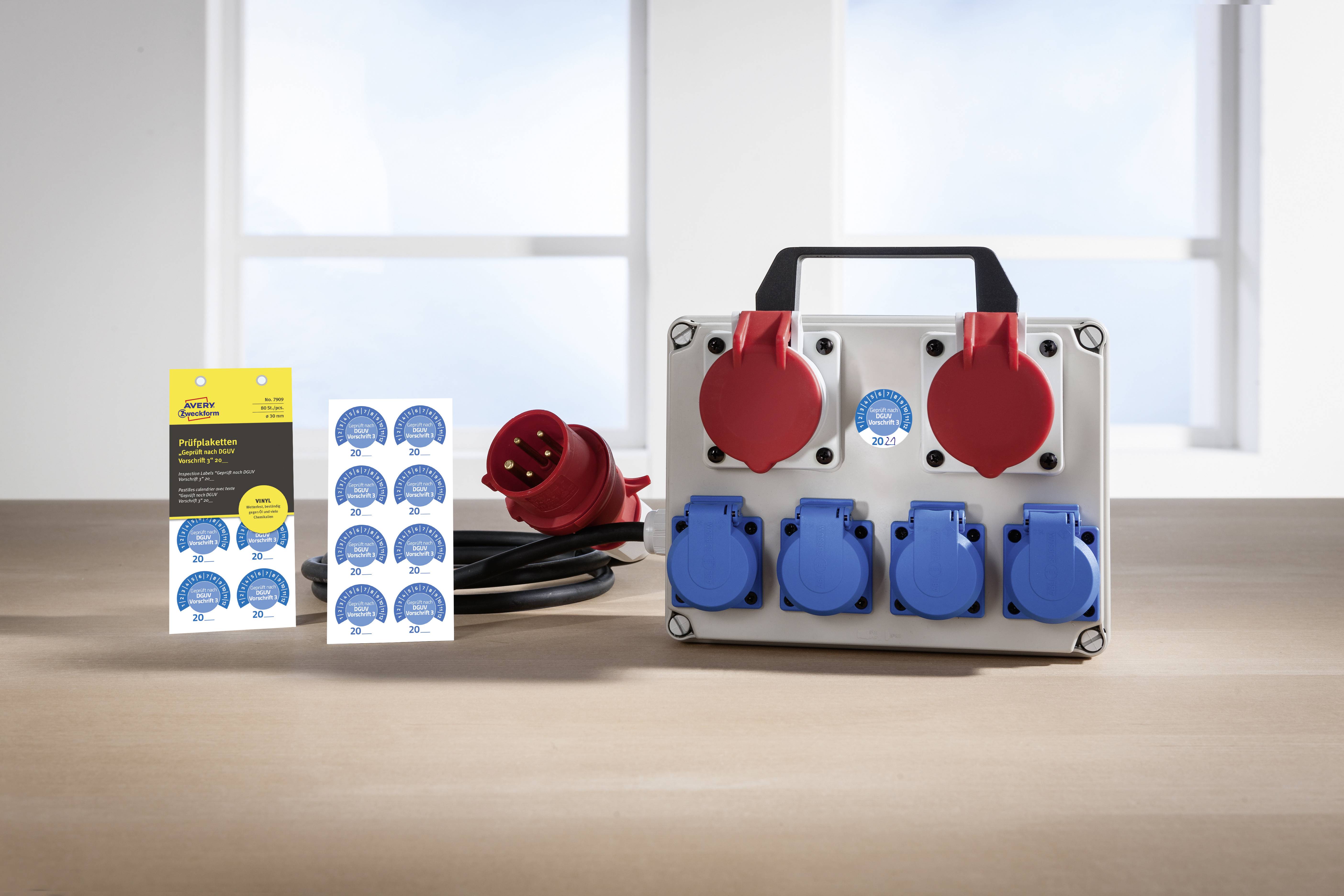 Ein tragbarer Stromverteiler mit blauen und roten Steckdosen auf einem Tisch, daneben eine gelbe Verpackung und ein rotes Kabel.