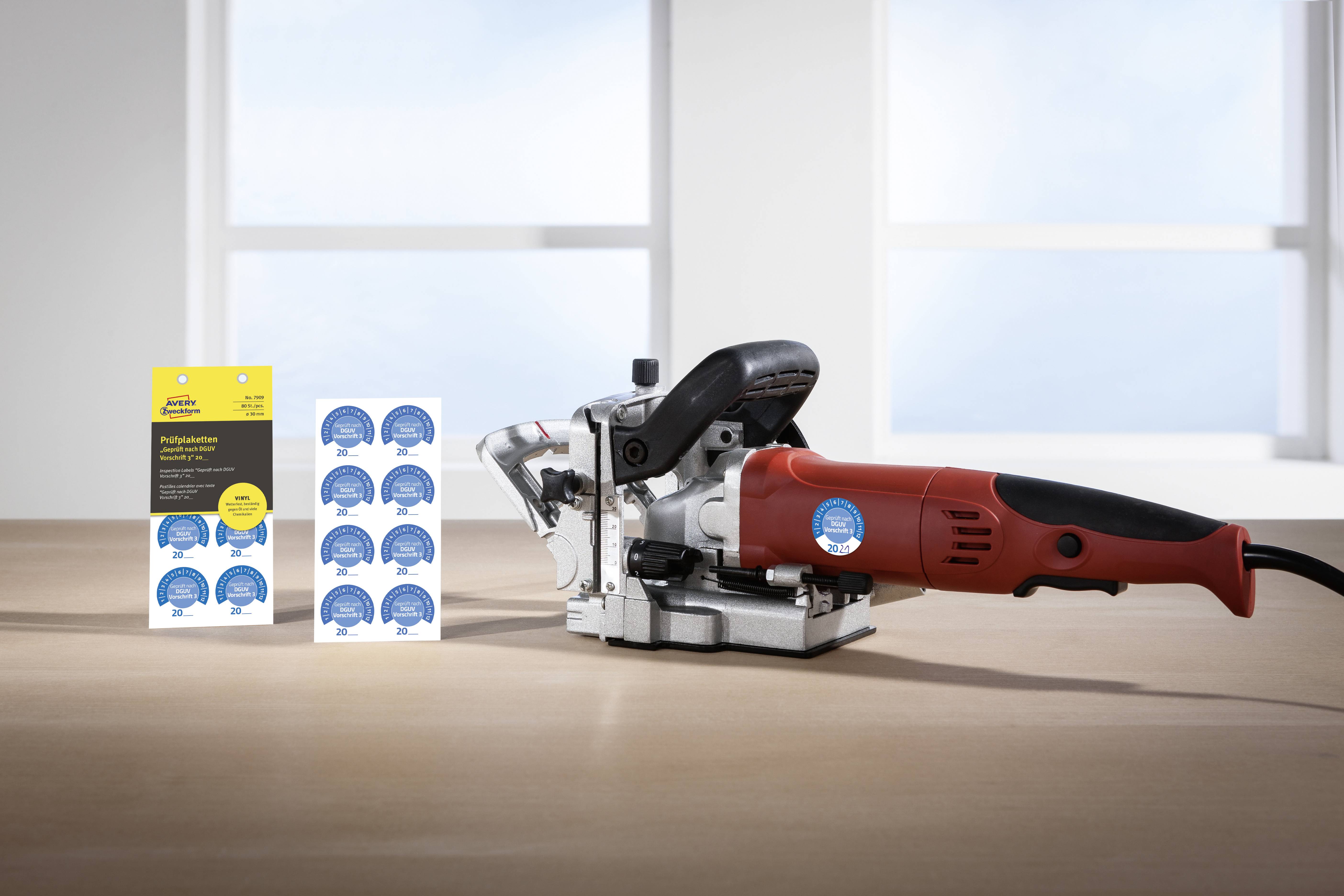 Ein rotes Elektrowerkzeug und ein Paket mit runden blauen Aufklebern auf einem Tisch vor einem Fenster.