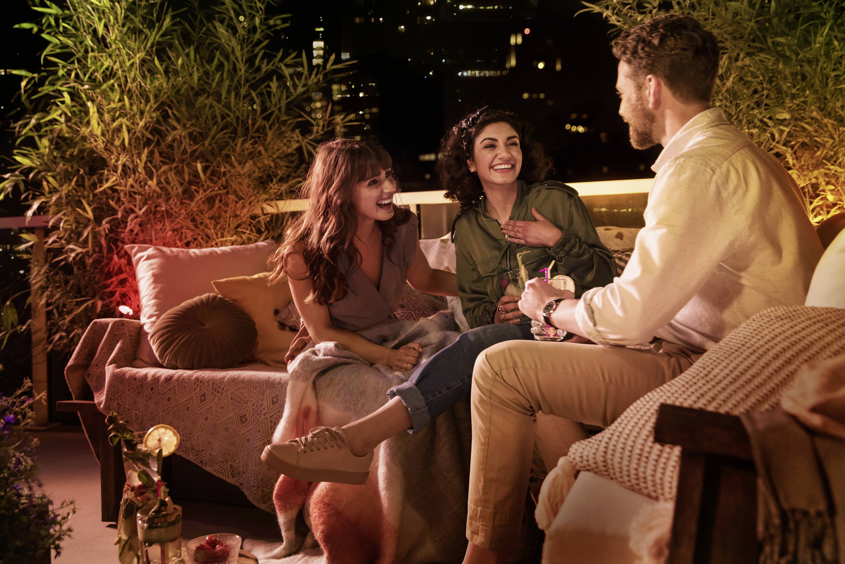 Drei Personen unterhalten sich lachend auf einer gemütlichen, beleuchteten Terrasse in der Nacht, umgeben von Pflanzen und Kissen.