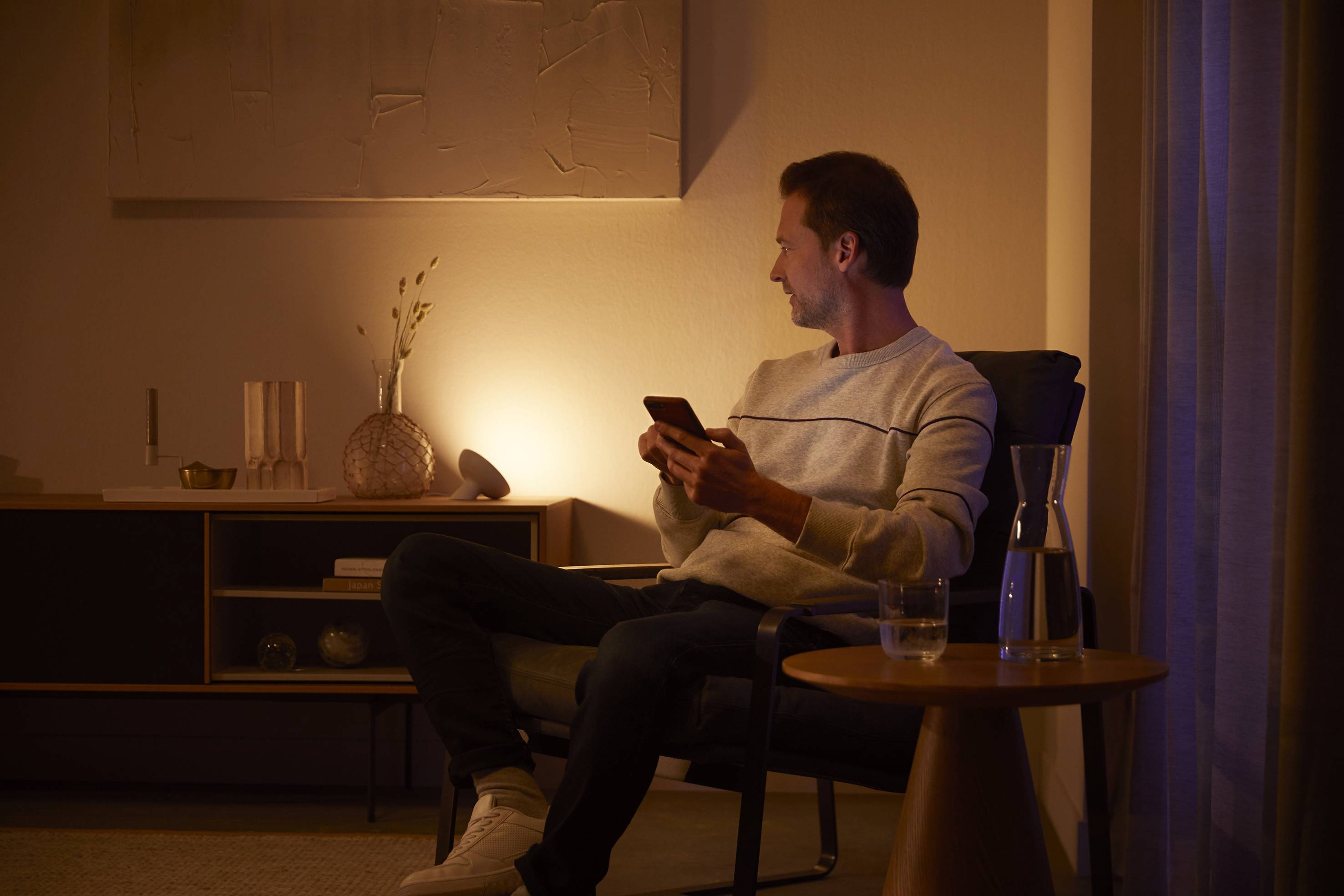 Ein Mann sitzt abends auf einem Stuhl in einem dimm beleuchteten Raum, hält ein Smartphone und schaut zur Seite. Im Hintergrund steht ein Tisch.