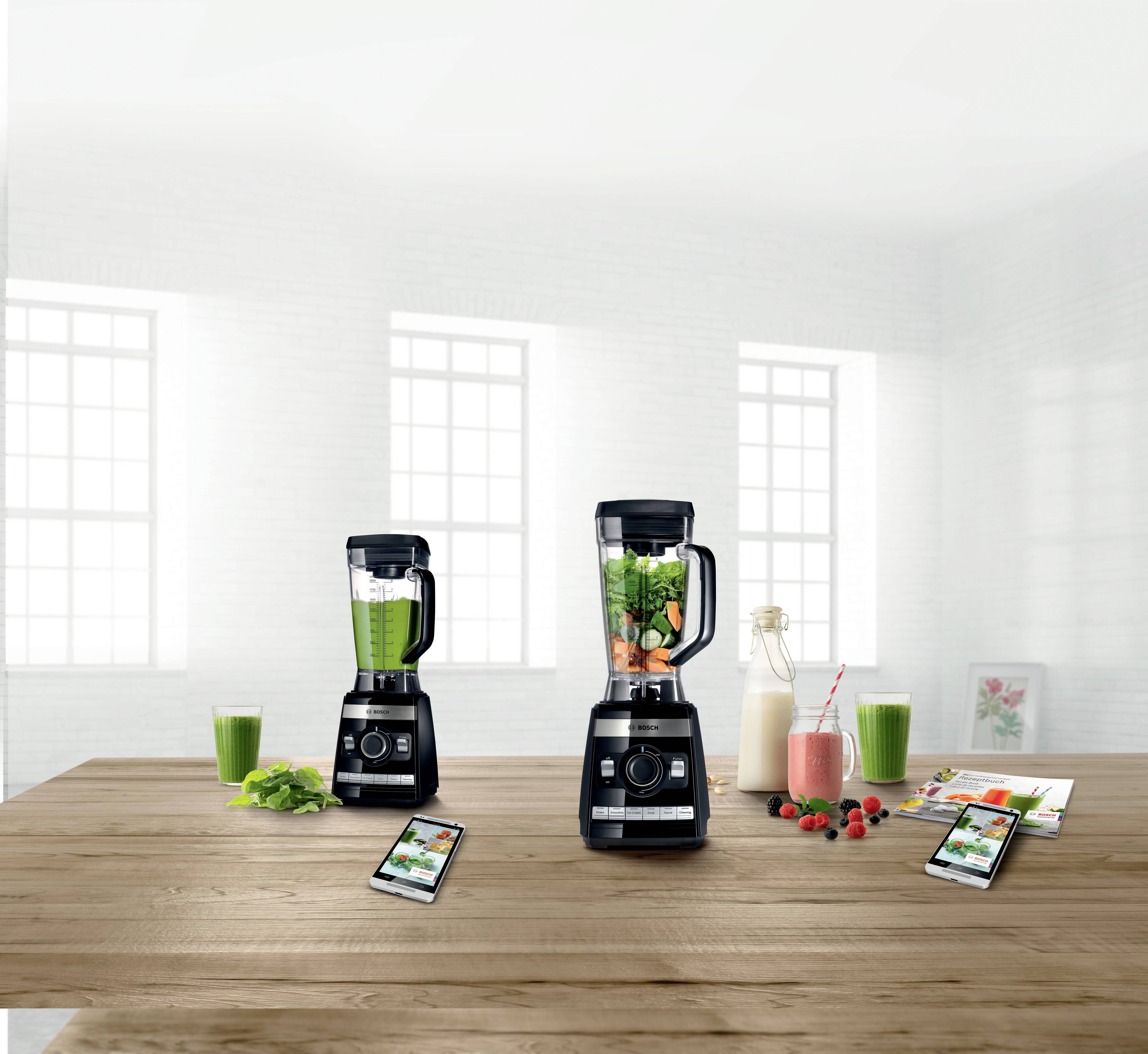 Zwei schwarze Mixer auf einem Holztisch, gefüllt mit grünen und bunten Zutaten. Daneben sind Smoothies in Gläsern sowie Getränke zu sehen.