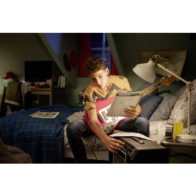 Ein Junge sitzt auf einem Bett, hält eine rote E-Gitarre und studiert Noten, während er einen Verstärker einstellt.