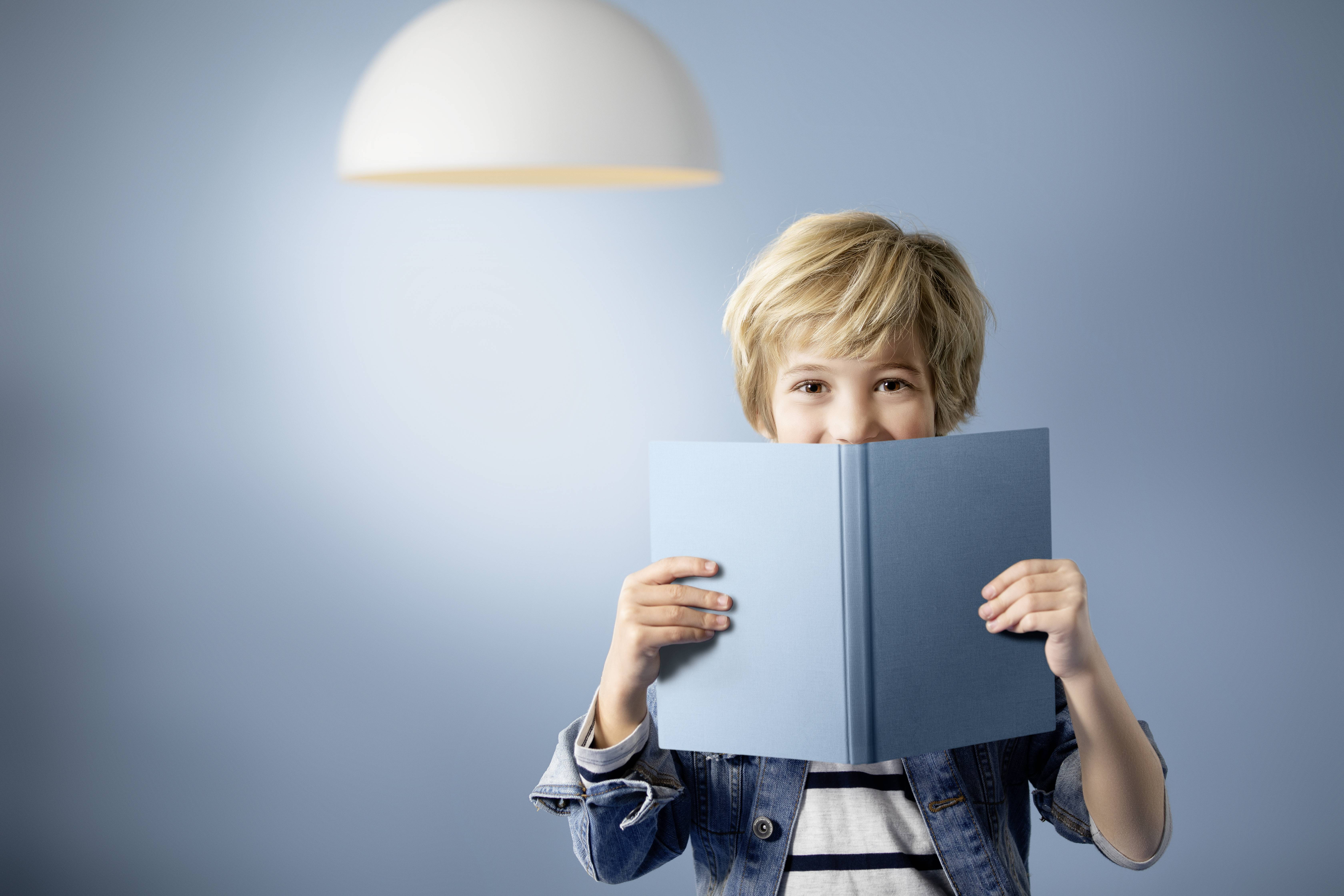 Ein Kind mit blonden Haaren hält ein blaues Buch und lächelt, während es in die Kamera schaut. Hintergrund und Buch sind in ähnlichen Blautönen.