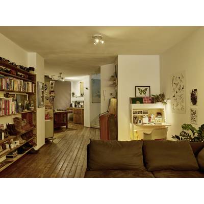 Wohnzimmer mit Holzboden, Bücherregal und Schreibtisch. Im Hintergrund eine Küche. Gemütliche Atmosphäre mit warmer Beleuchtung.