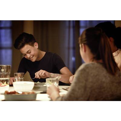 Ein Teenager sitzt lächelnd an einem Esstisch mit anderen Personen, isst eine Mahlzeit. Die Szene wirkt freundlich und gemütlich.