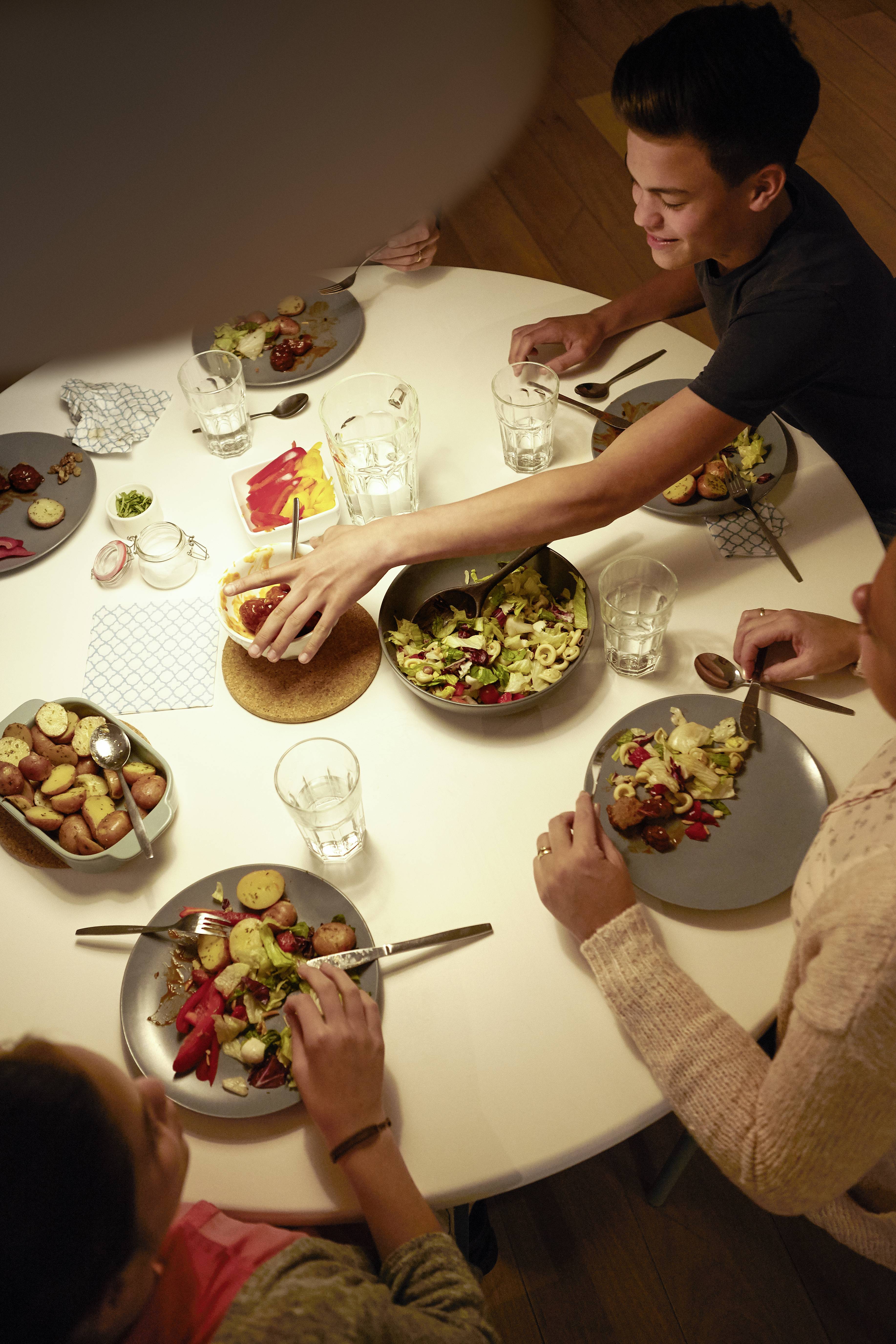 Menschen essen gemeinsam an einem runden Tisch. Verschiedene Gerichte wie Salat und Kartoffeln sind serviert, eine Person reicht Essen weiter.