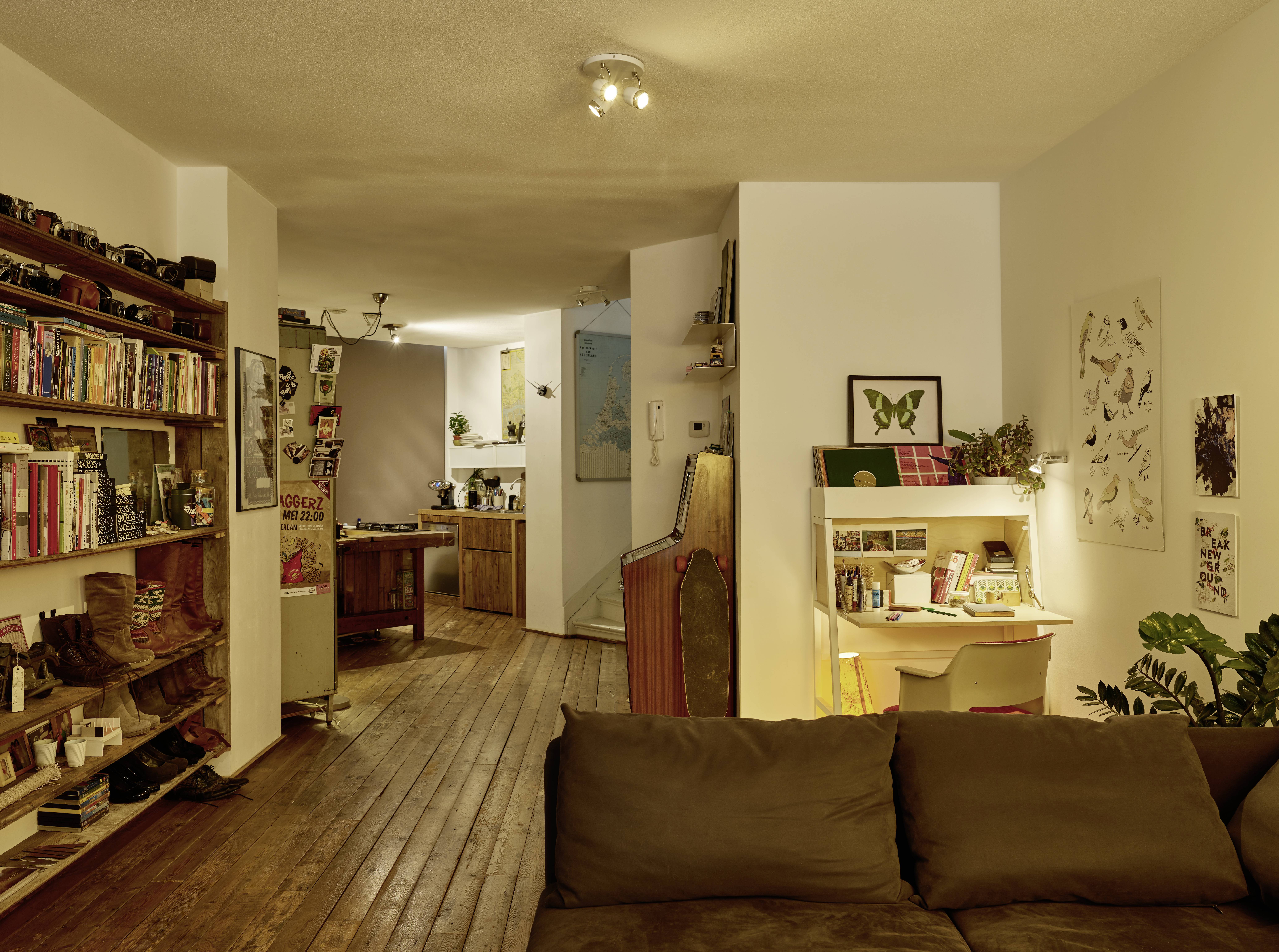 Wohnzimmer mit Holzboden, Bücherregalen und Schreibtischecke. Im Hintergrund eine offene Küche. Gemütliches und einladendes Ambiente.