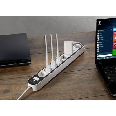 Eine Steckdosenleiste mit mehreren Steckdosen und eingesteckten Geräten auf einem Holztisch neben einem eingeschalteten Laptop.