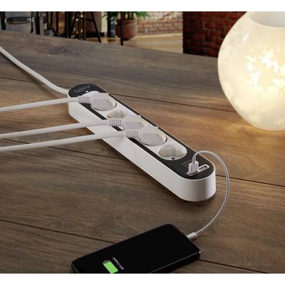 Ein Ladegerät mit mehreren Steckdosen und USB-Anschluss lädt ein Smartphone, das auf einem Tisch neben einer dekorativen Lampe liegt.