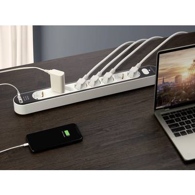 Mehrfachsteckdose auf einem Holztisch mit fünf eingesteckten Kabeln, Smartphone lädt, Laptop daneben eingeschaltet.