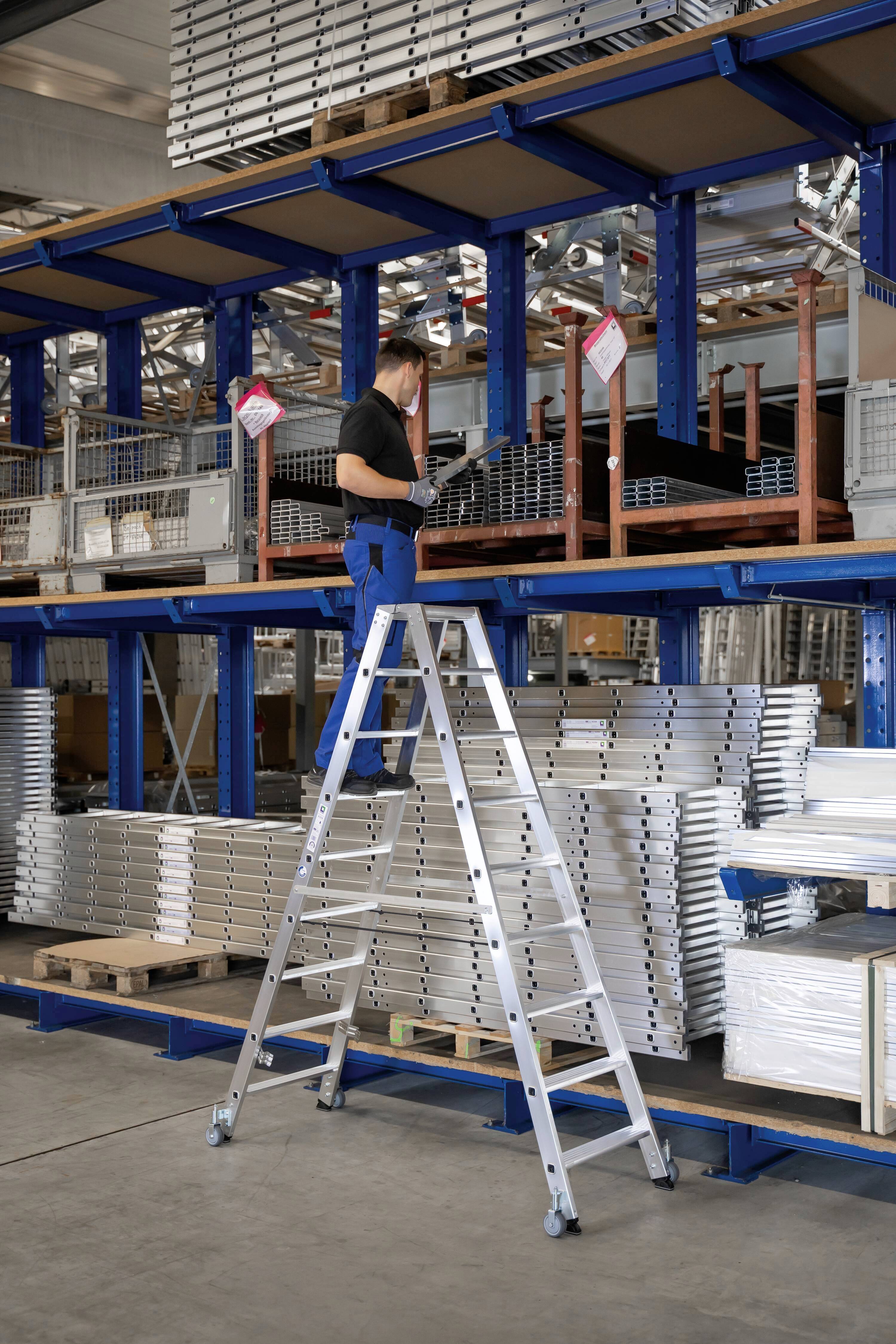 Eine Person steht auf einer Leiter in einem Lager, erreicht ein Regal mit Aluminiumprofilen.