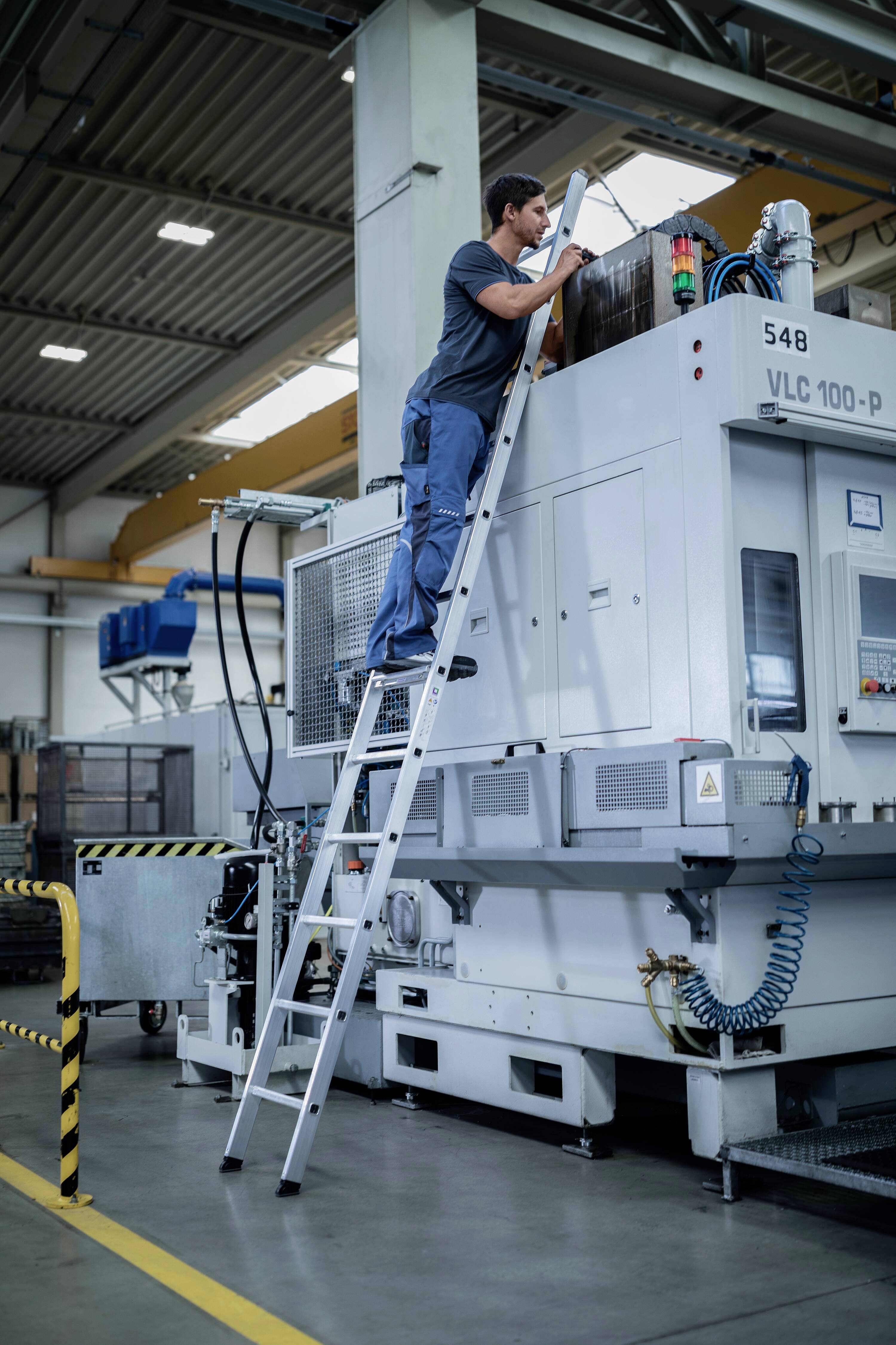 Ein Mann steht auf einer Leiter und arbeitet an einer großen grauen Maschine in einer Fabrikumgebung. Die Maschine hat Beschriftungen und Schaltungen.
