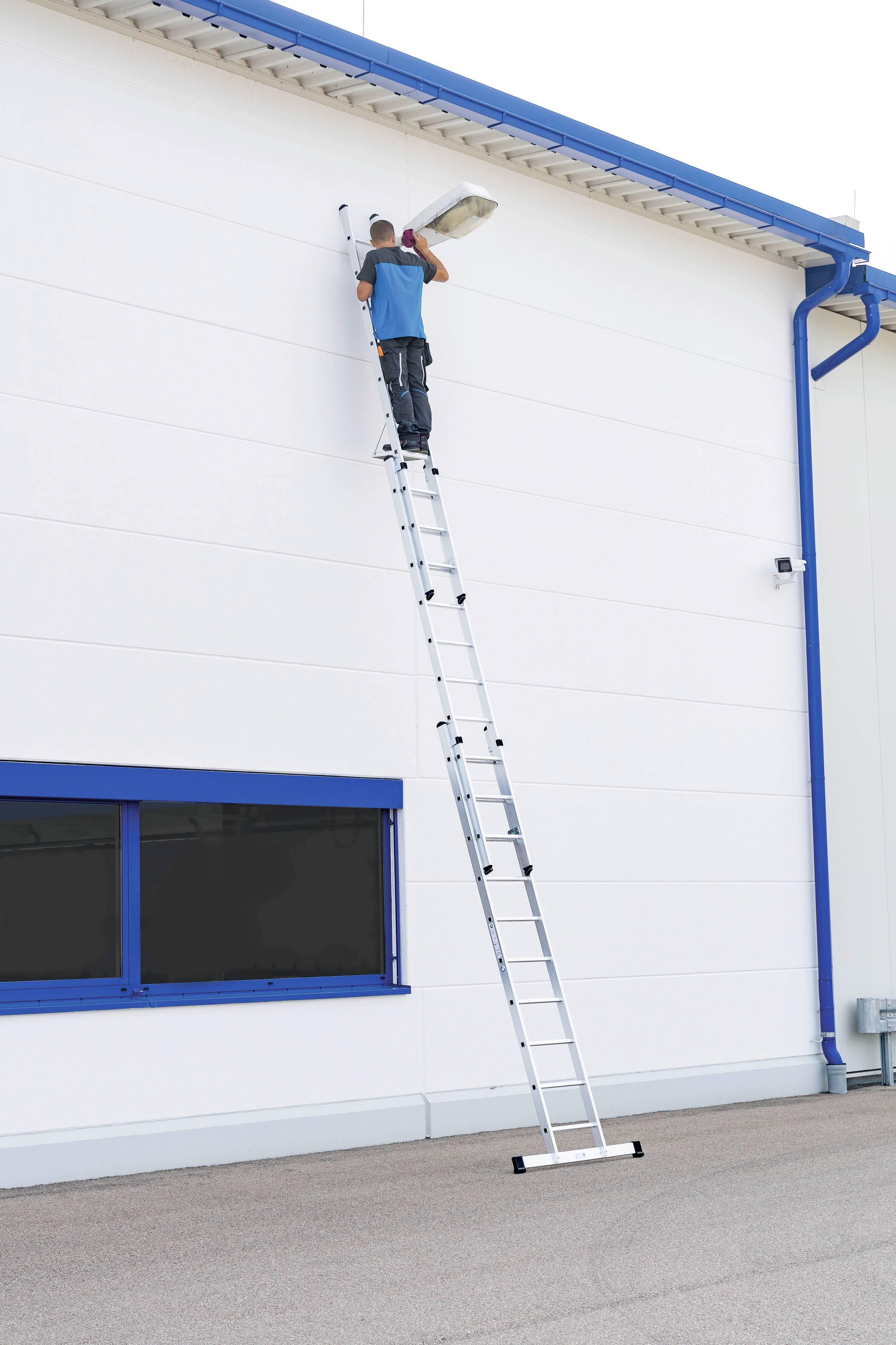 Eine Person auf einer langen Leiter tauscht an einer weißen Gebäudewand eine Außenbeleuchtung aus, unterhalb ein Fenster mit blauem Rahmen.