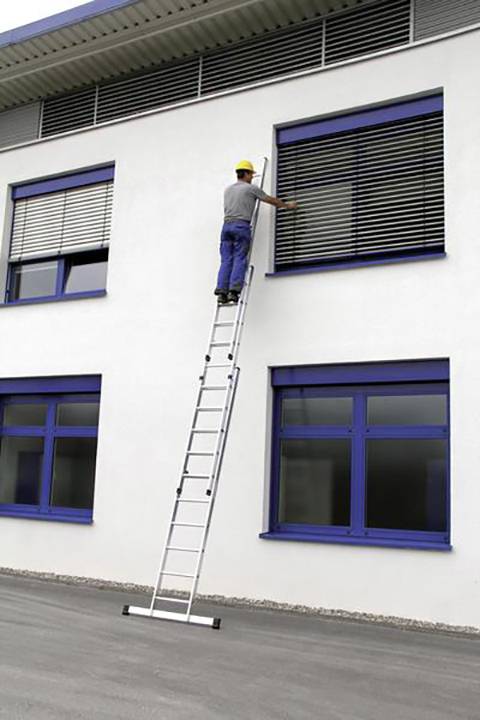 Ein Bauarbeiter in blauer Jeans und gelbem Helm steht auf einer Leiter und repariert ein Fenster an einem weiß gestrichenen Gebäude.