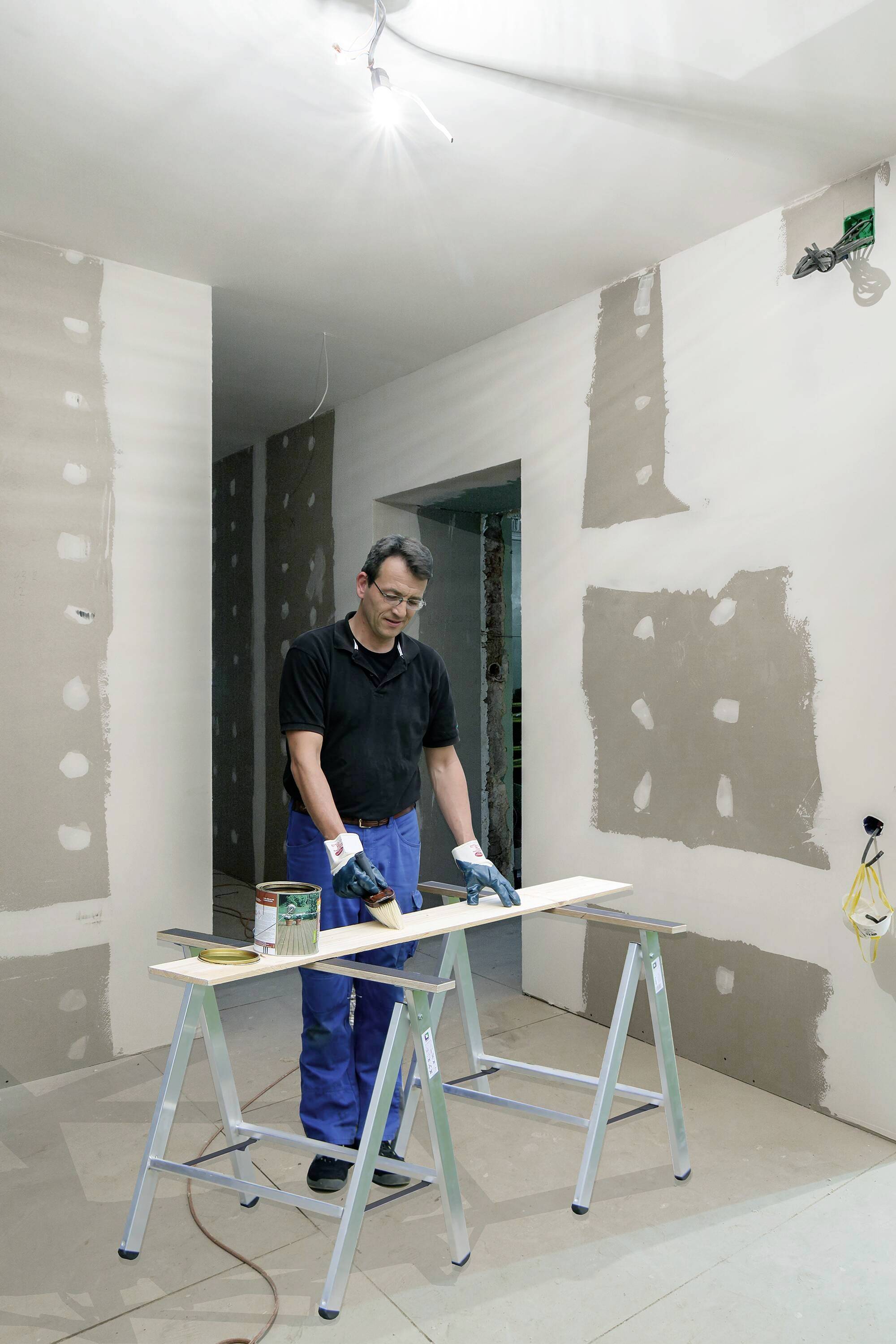 Ein Mann im schwarzen Shirt und blauen Hosen streicht ein Holzbrett in einem Raum mit unvollendeten Trockenbauwänden.