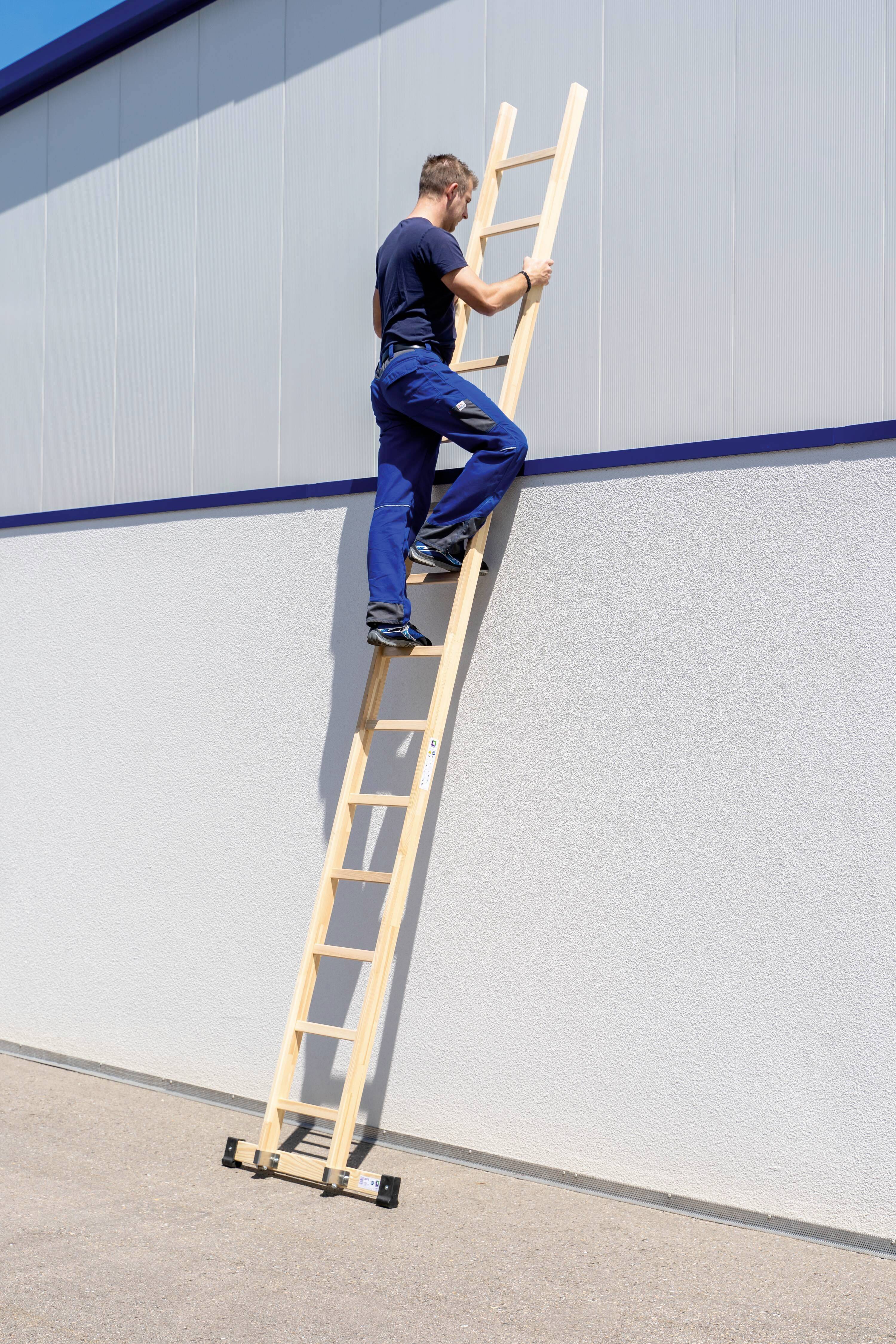 Ein Mann steigt eine Holzleiter hinauf, die an einer großen, grauen Hauswand lehnt. Der Himmel ist blau, und es ist sonnig.