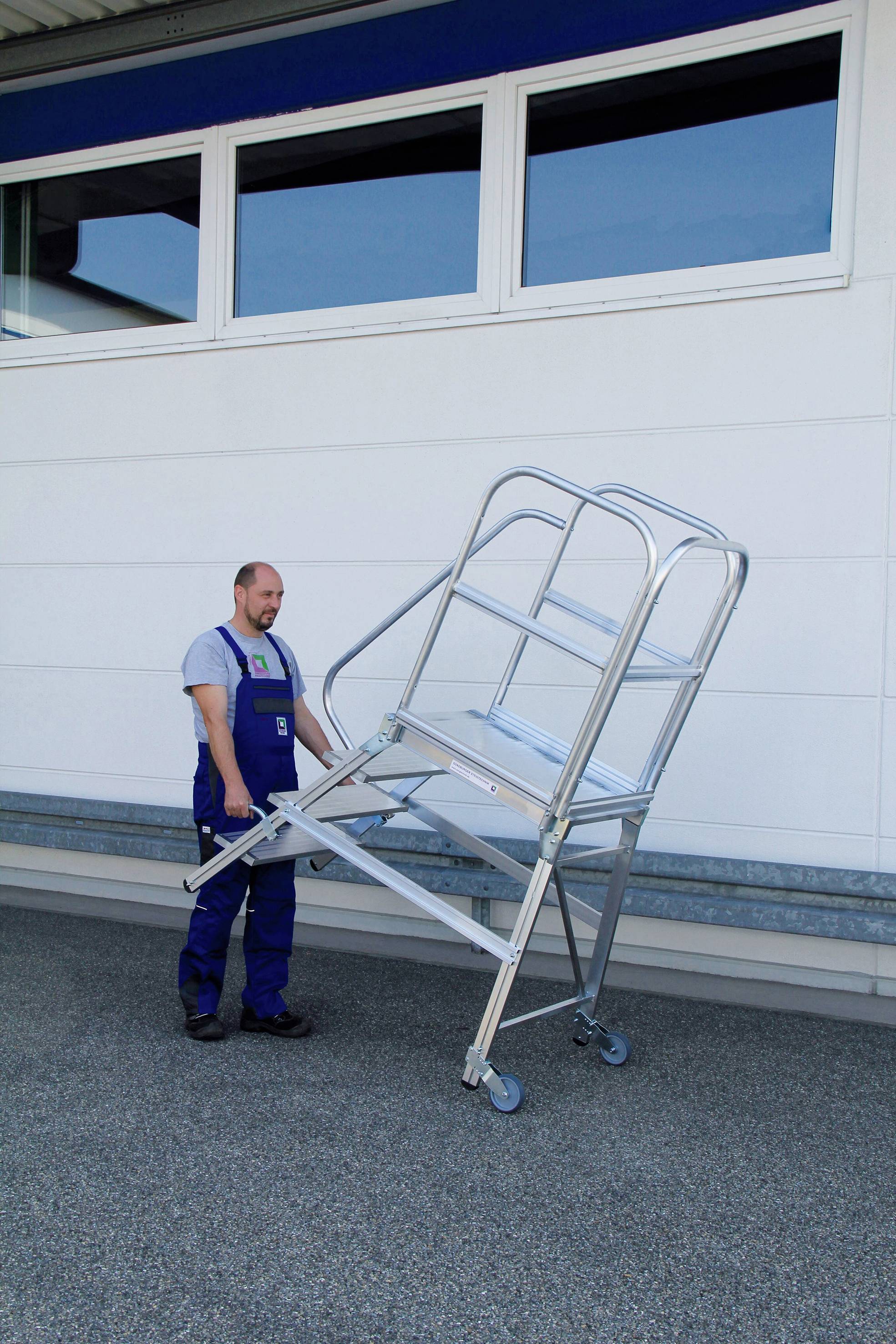 Ein Mann in Arbeitskleidung bewegt eine mobile Arbeitstreppe mit Rollen vor einem Gebäude. Die Treppe hat ein Geländer für Sicherheit.