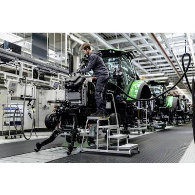 Ein Arbeiter montiert Teile an einem Traktor in einer Produktionshalle. Mehrere Traktoren in verschiedenen Bauphasen sind sichtbar.