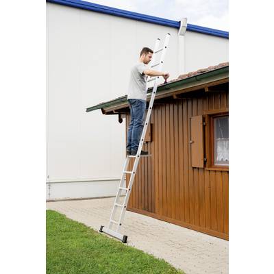 Ein Mann steht auf einer Leiter und repariert das Dach eines kleinen Holzhauses neben einer weißen Gebäudewand.