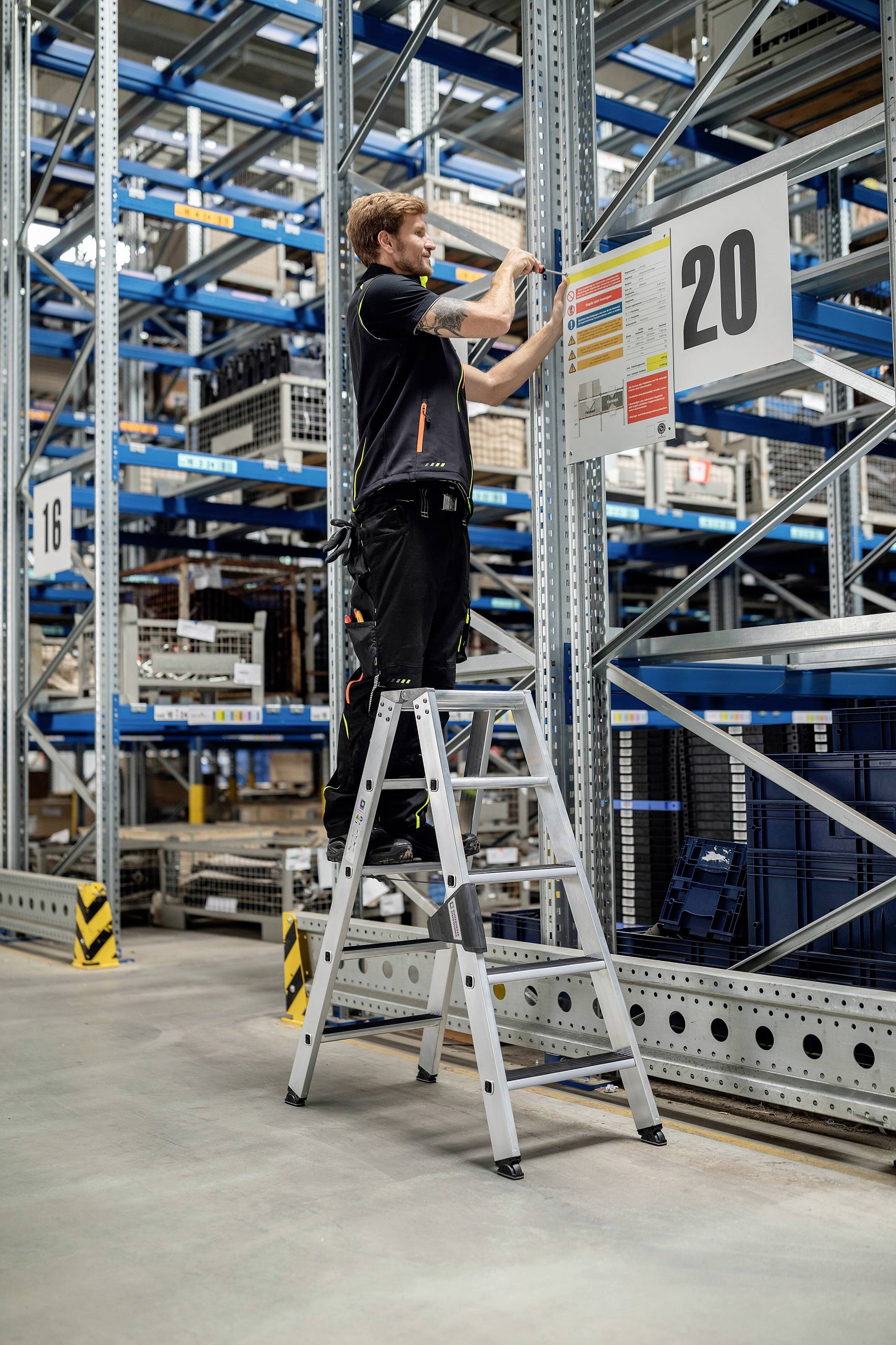 'Ein Arbeiter steht auf einer Leiter in einem Lager und befestigt ein großes Schild mit der Nummer 20 an einem Metallregal.'