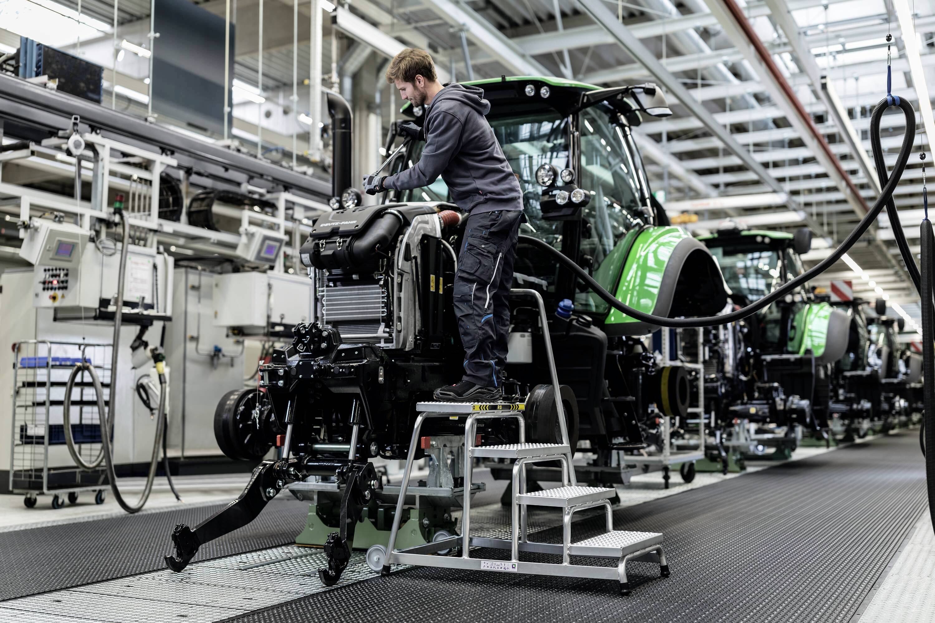 Ein Arbeiter montiert einen Traktor in einer Fabrikhalle. Mehrere grüne Traktoren sind in einer Produktionslinie zu sehen.