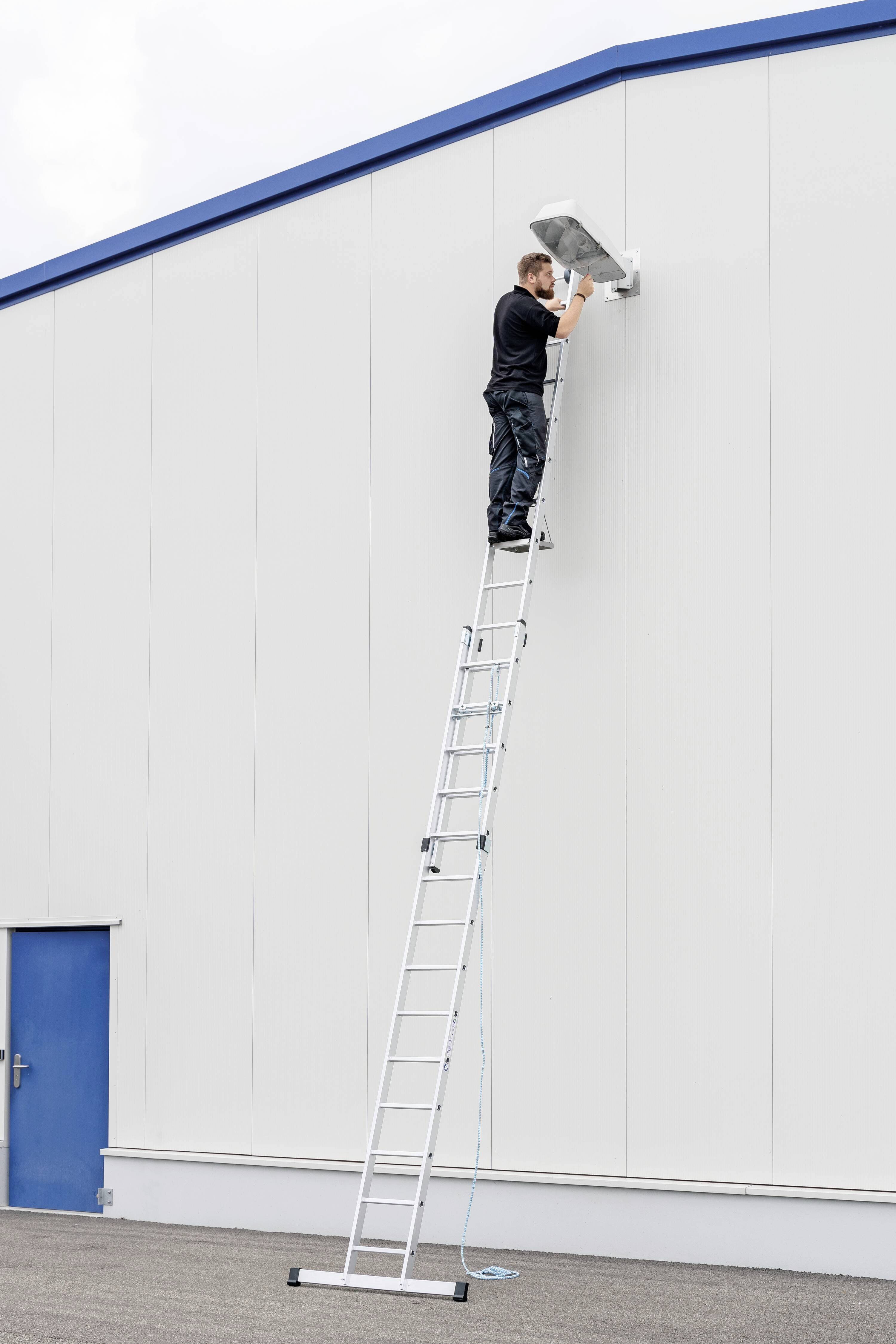 Ein Mann steht auf einer langen Leiter und arbeitet an einer Überwachungskamera an der Wand eines Gebäudes.