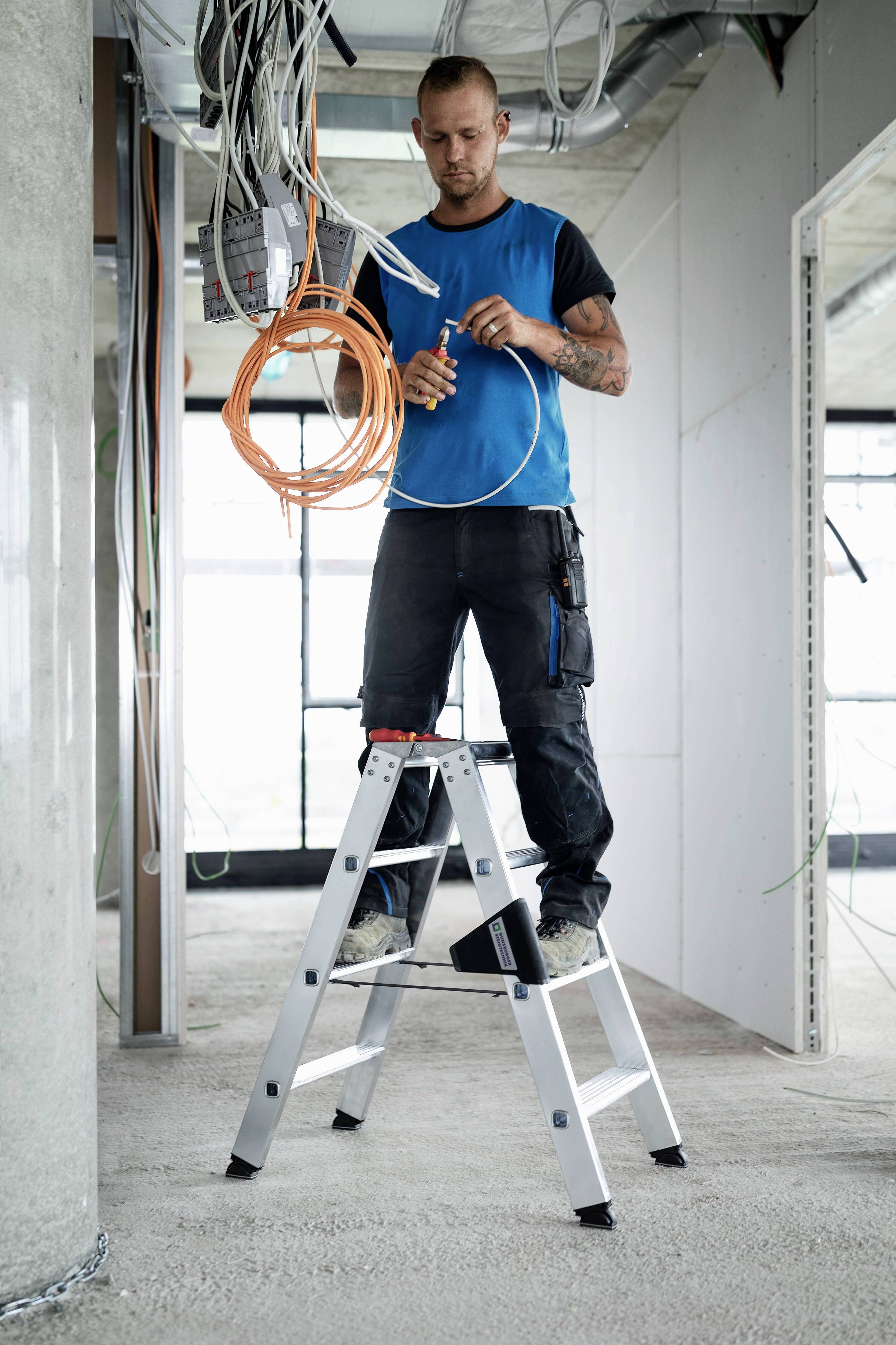 Ein Mann steht auf einer Leiter in einem halbfertigen Raum und arbeitet mit Kabeln, die von der Decke hängen.