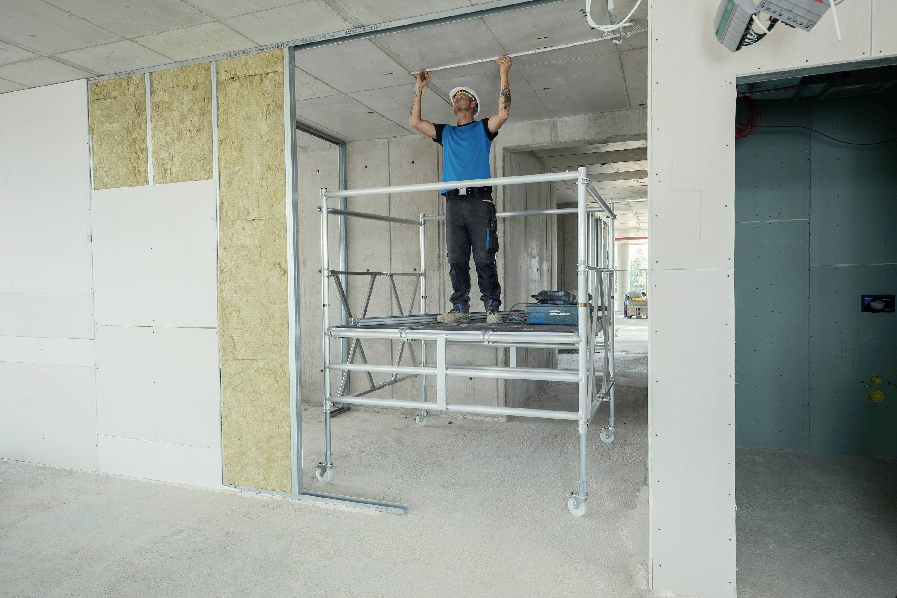 Ein Mann steht auf einem rollbaren Gerüst in einem unfertigen Raum, verwendet Werkzeuge zur Installation von Deckenbeleuchtung.