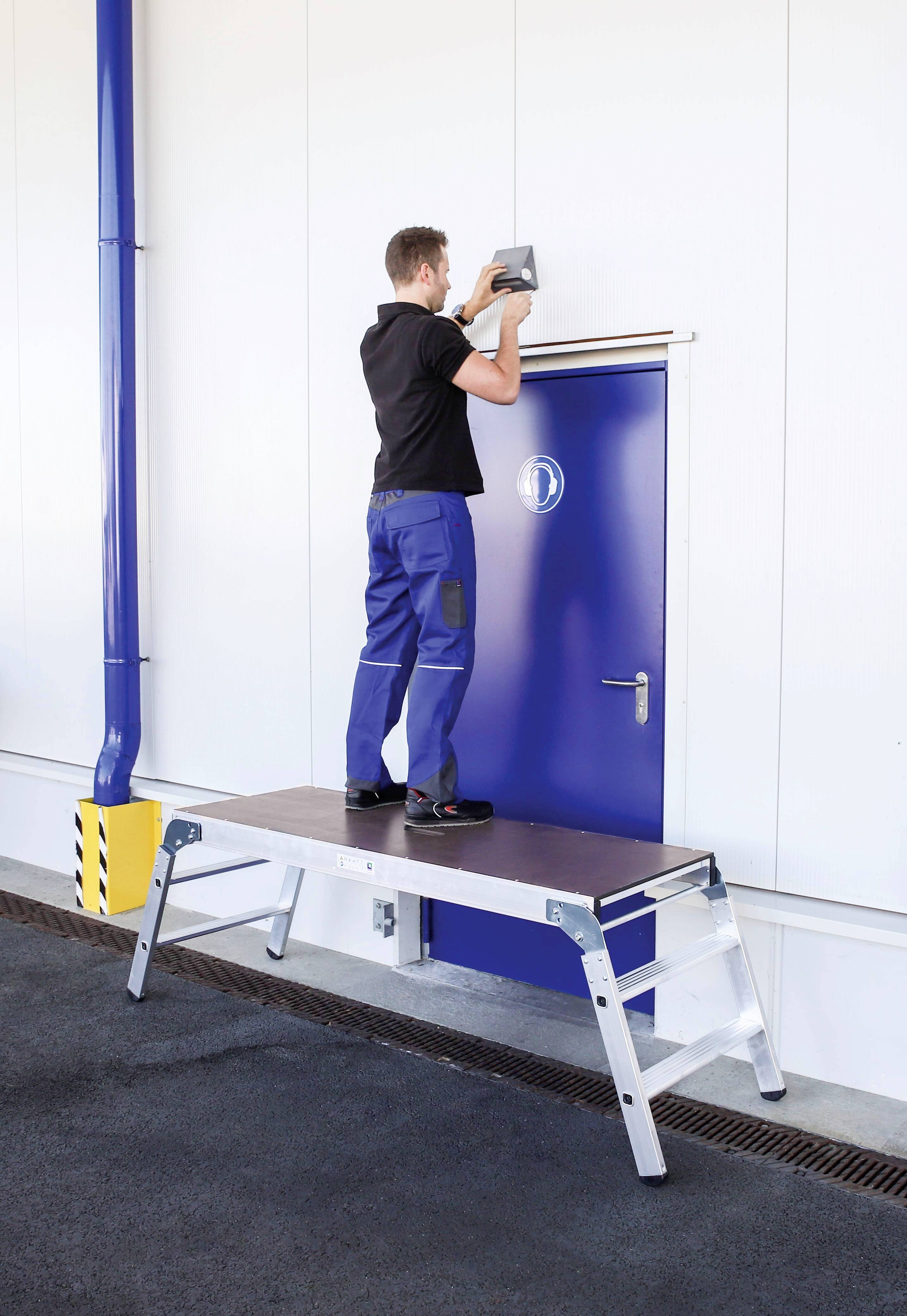 Ein Mann steht auf einer Plattform zwischen zwei Leitern und montiert eine Vorrichtung über einer blauen Tür in einer industriellen Umgebung.