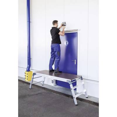 Ein Mann steht auf einer stabilen Plattformleiter und arbeitet an einer Vorrichtung über einer blauen Tür in einem industriellen Umfeld.