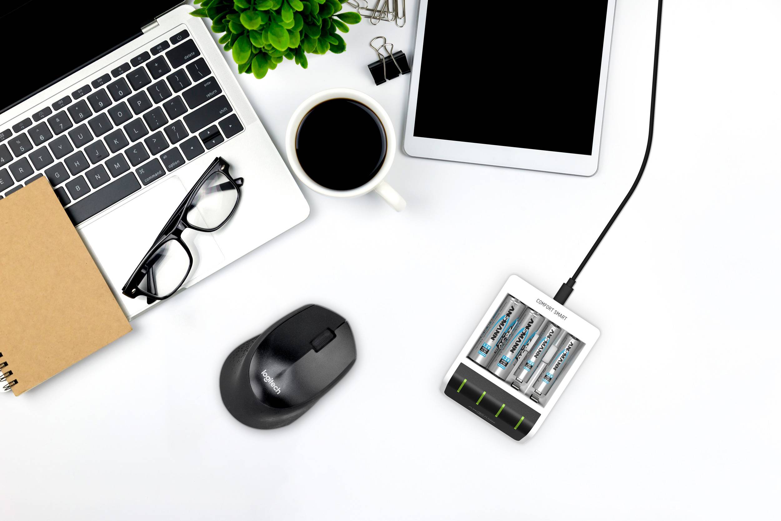 Ein Schreibtisch mit Laptop, Tablet, Brille, Kaffeetasse, kabelloser Maus und Batterieladegerät. Eine Pflanze verleiht etwas Grün.