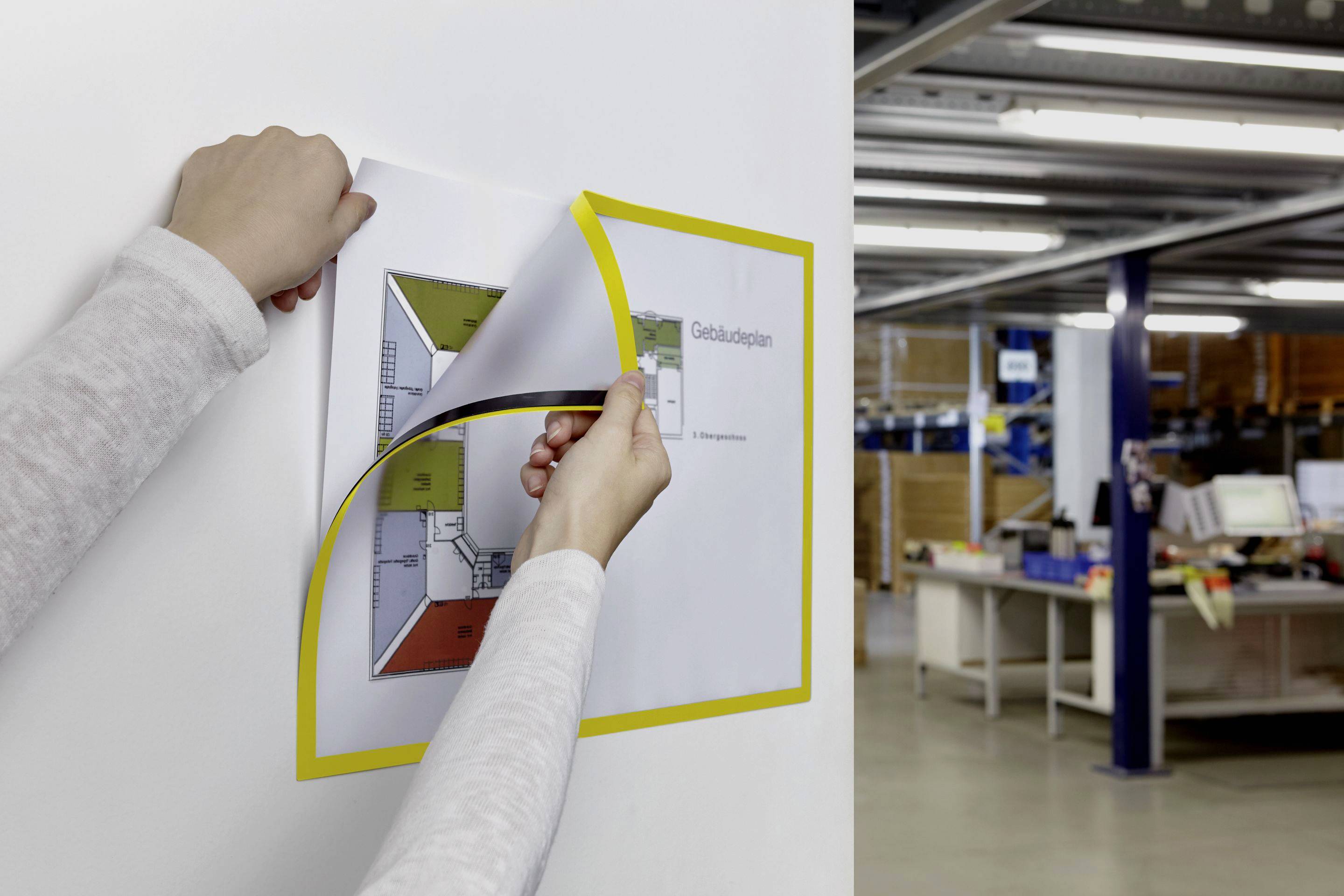 Eine Person befestigt einen Gebäudeplan an einer Wand in einem Büro oder Lager. Im Hintergrund sind Regale und Büroschreibtische zu sehen.