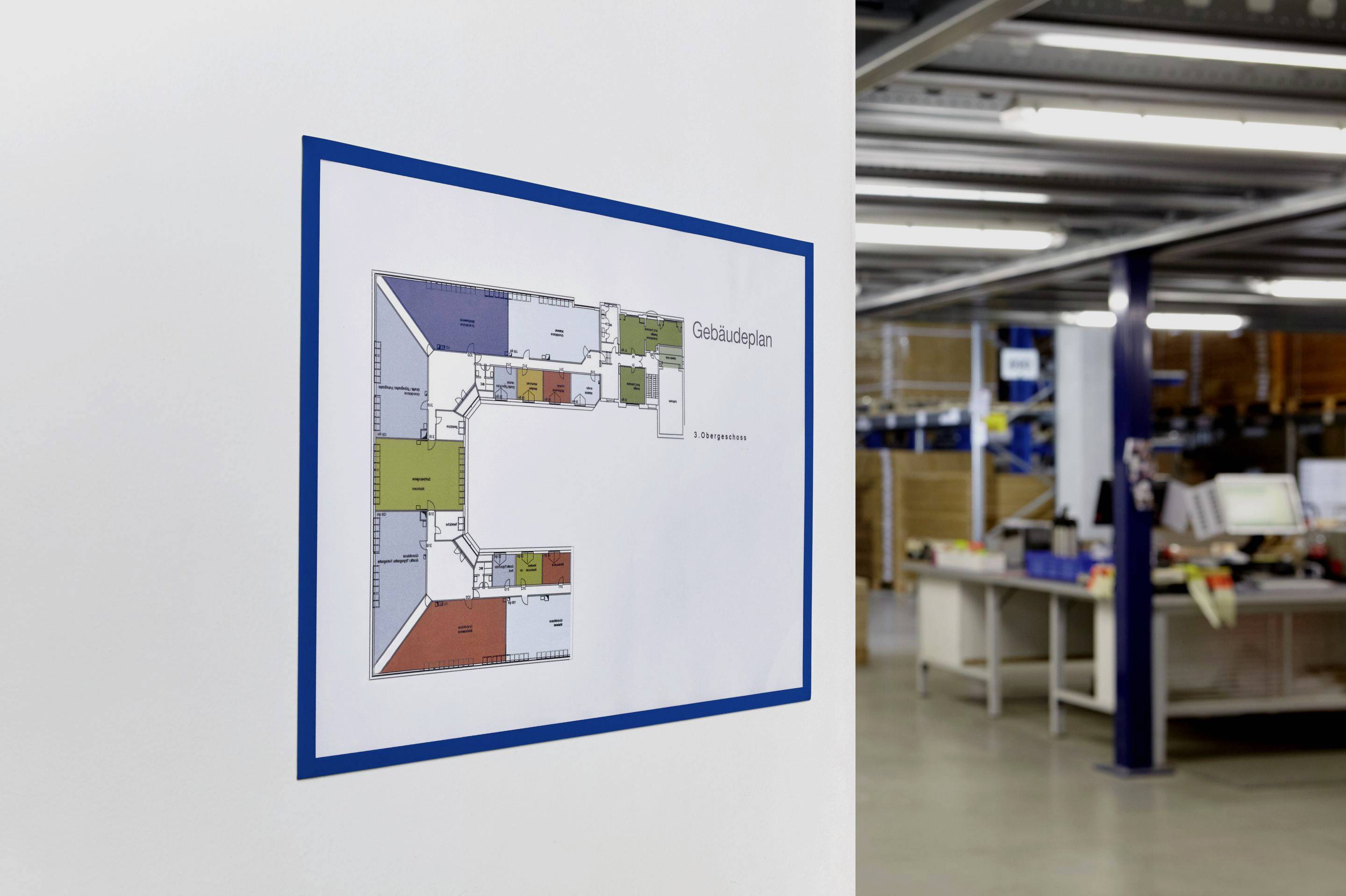 Gebäudeplan an der Wand eines Lagers, zeigt farbige Räume und Gänge. Büro- und Arbeitsbereich im Hintergrund.